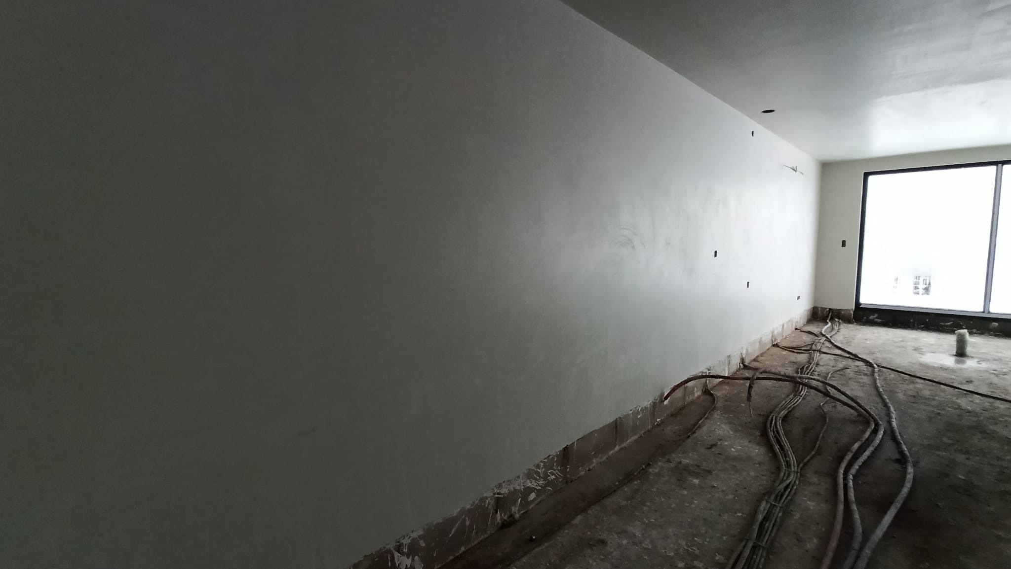 Świeżo położony, jasnoszary tynk na ścianie wewnętrznej pomieszczenia w trakcie wykańczania, widoczne kable elektryczne na podłodze wzdłuż ściany.