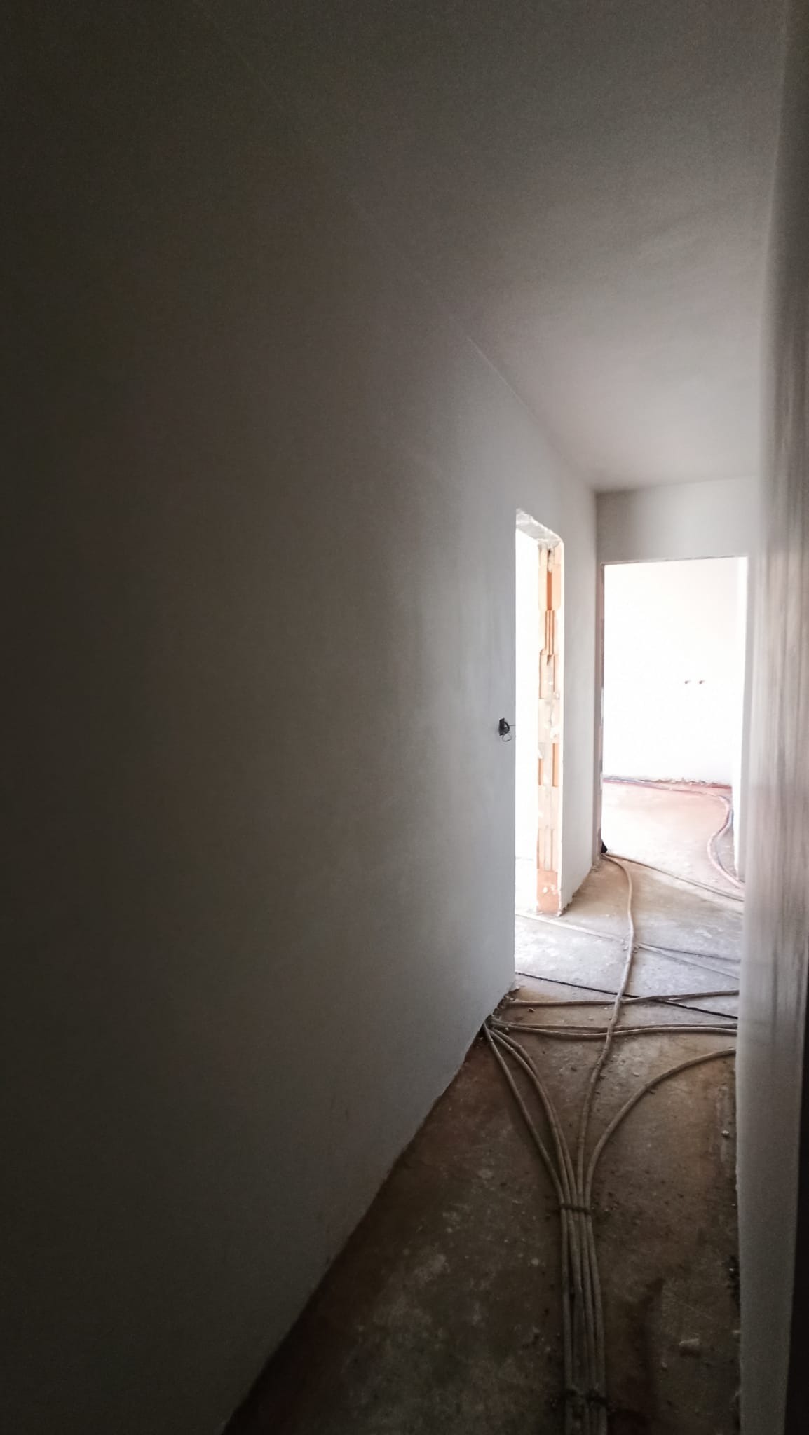 Świeżo otynkowane ściany korytarza wewnątrz budynku, z widocznymi przewodami elektrycznymi na podłodze i otwartymi drzwiami w tle.