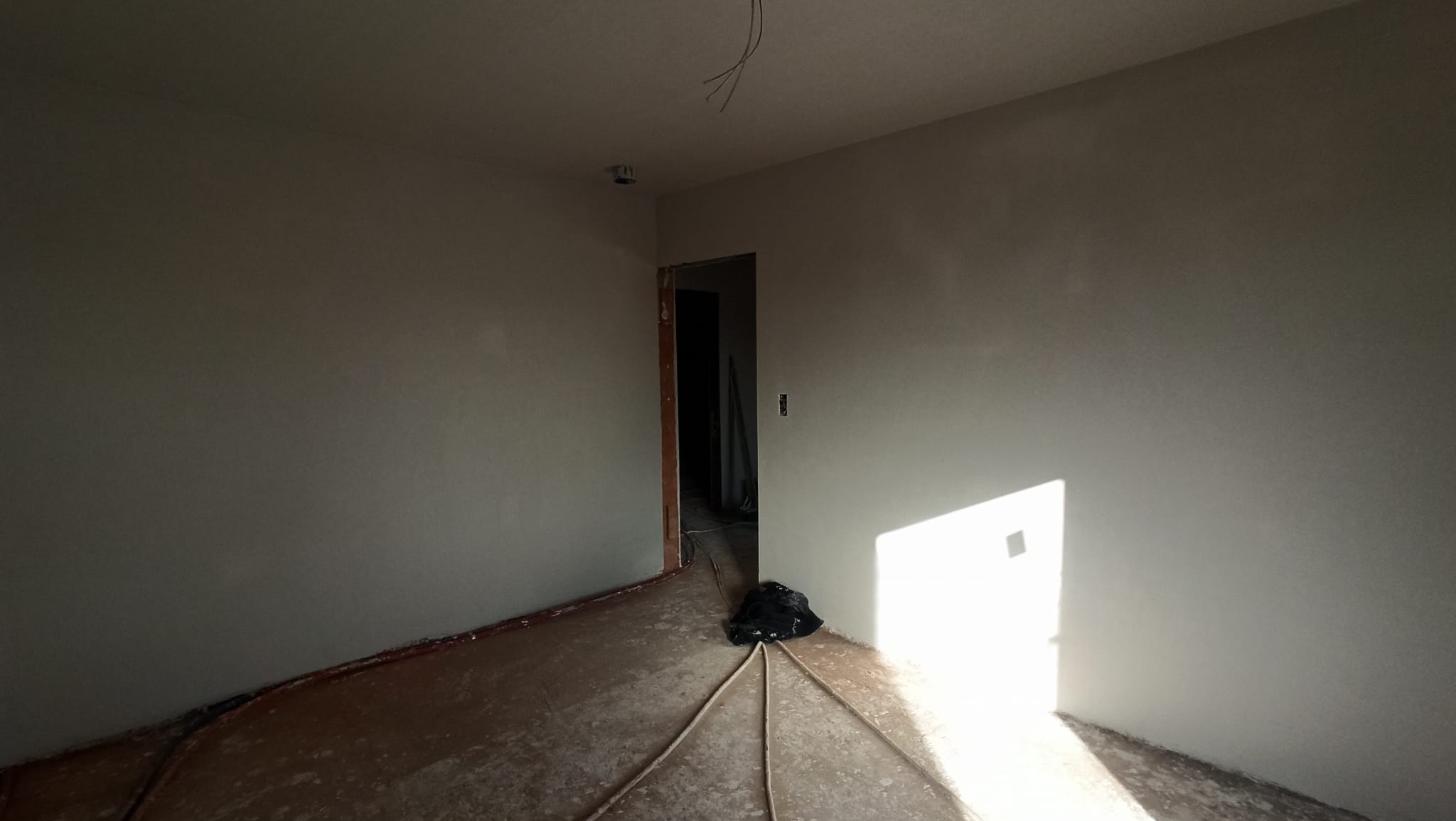 Wnętrze pomieszczenia po nałożeniu świeżych, jasnych tynków maszynowych, z widocznymi przewodami elektrycznymi wystającymi z sufitu i plamami na podłodze, oświetlone silnym promieniem słonecznym...