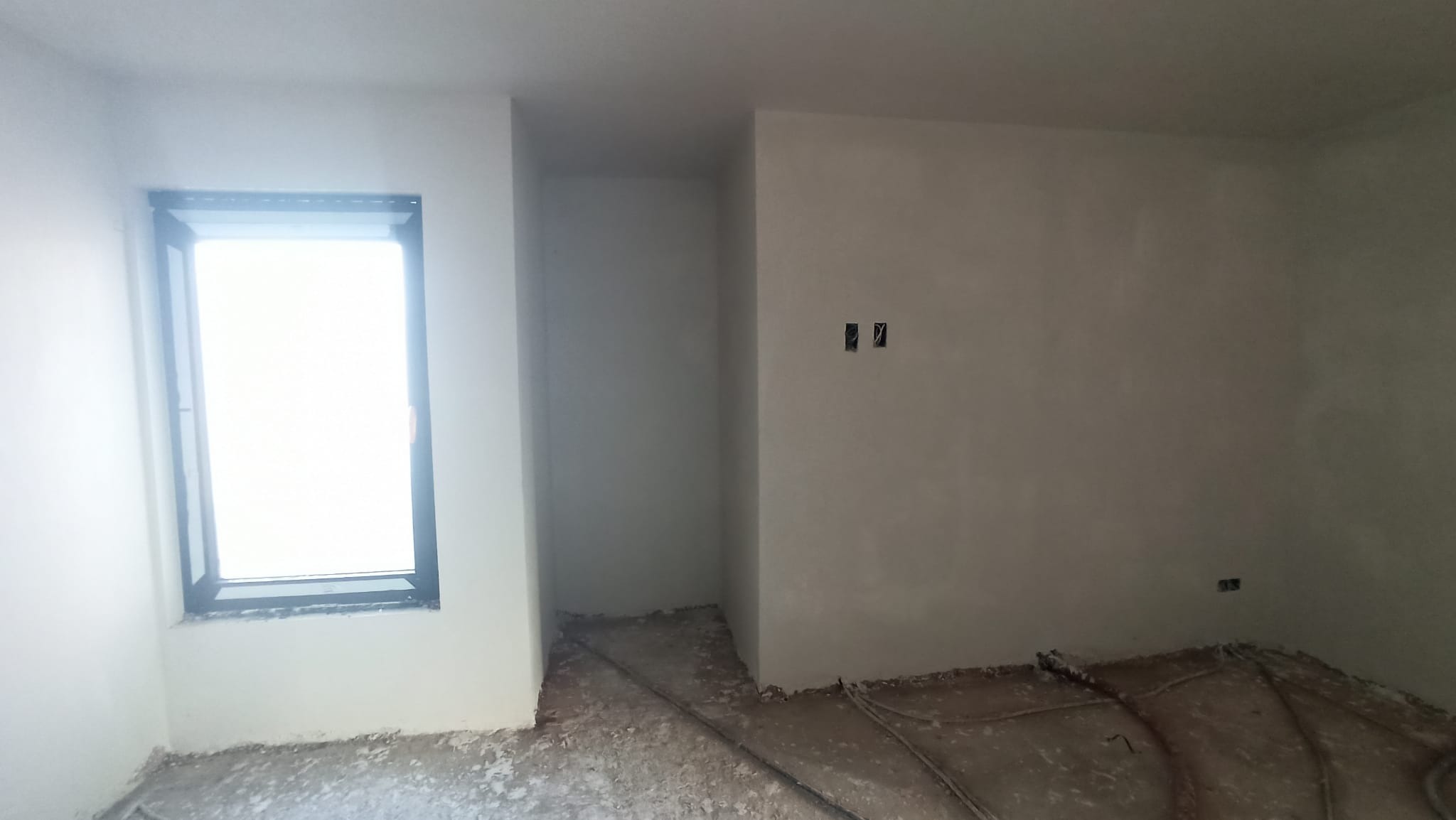 Wnętrze pokoju po nałożeniu gładzi gipsowej, widoczne gniazdka elektryczne i fragment okna z czarną ramą. Na podłodze widoczne ślady po pracach wykończeniowych.