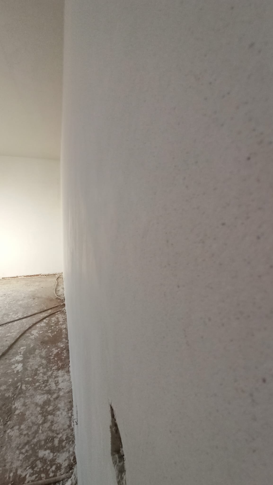 Świeżo nałożony tynk na ścianie wewnętrznej z widocznym ubytkiem w dolnej części, na podłodze resztki materiałów budowlanych i przewód.