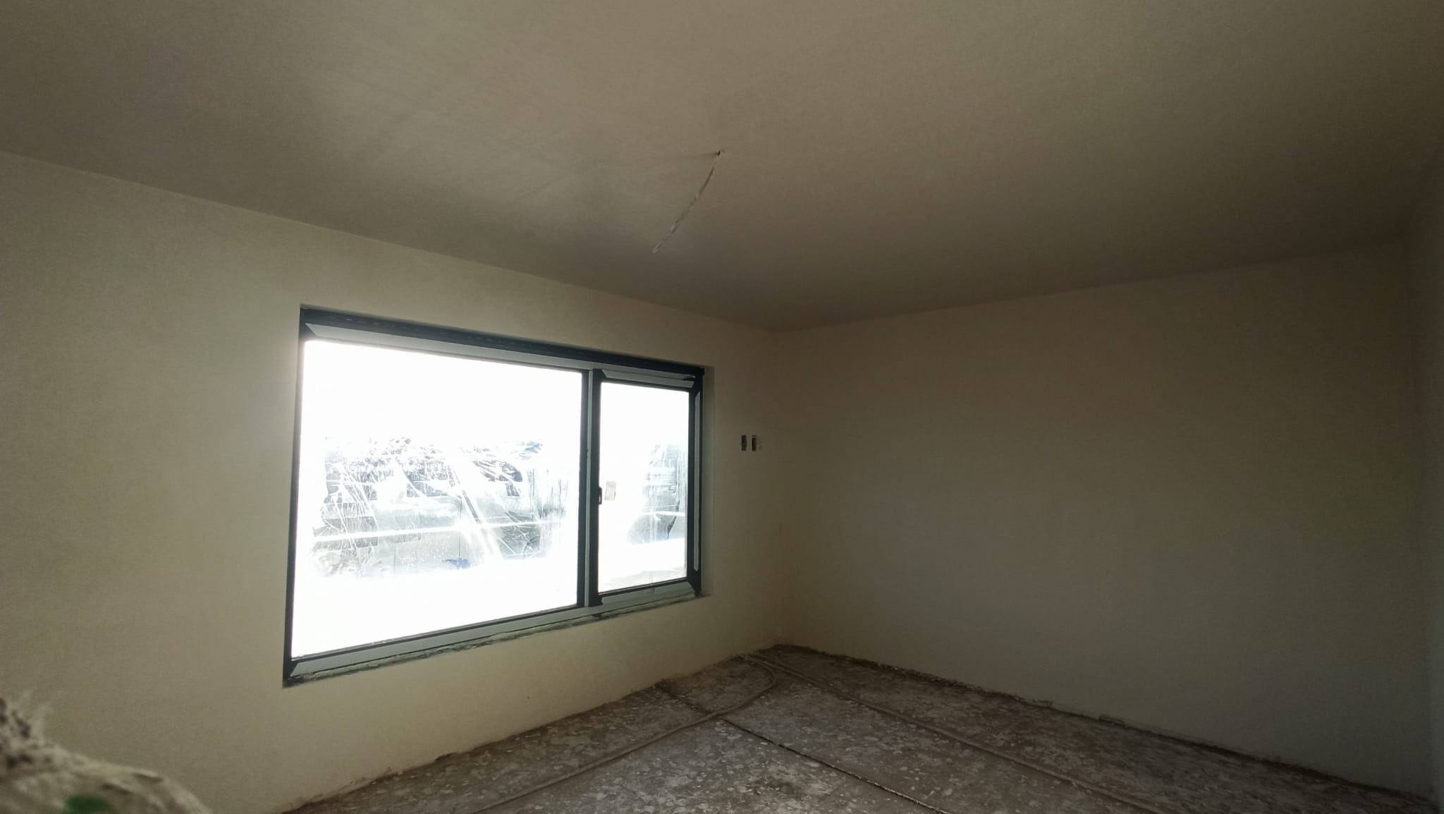 Pokój po nałożeniu jasnych tynków maszynowych, widoczne okno z zabezpieczeniem folią, szare rurki instalacyjne na podłodze.