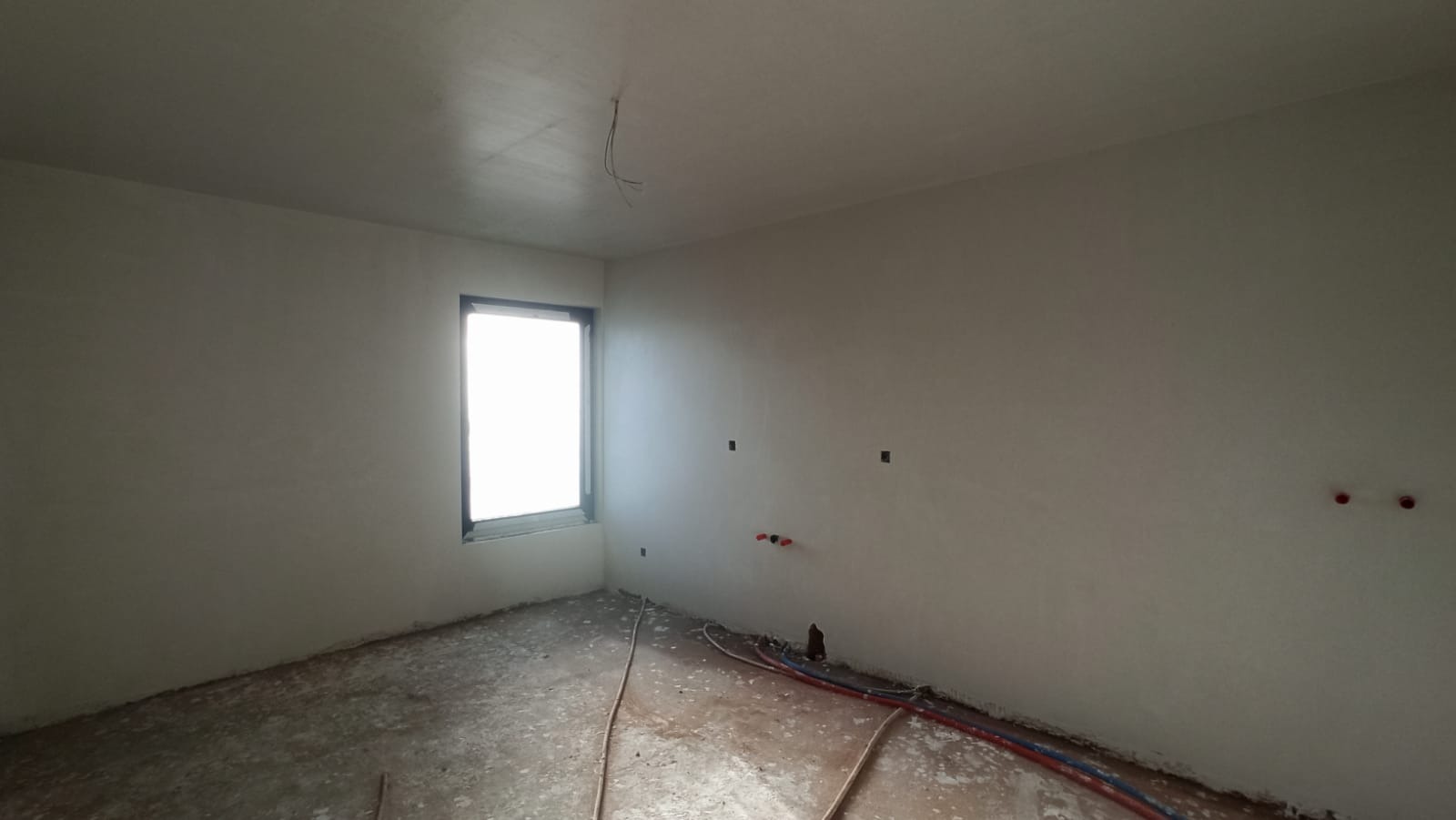 Wnętrze pokoju po nałożeniu gładzi gipsowej, widoczne instalacje wodne i elektryczne, surowy beton na podłodze, okno z widokiem na zewnątrz.