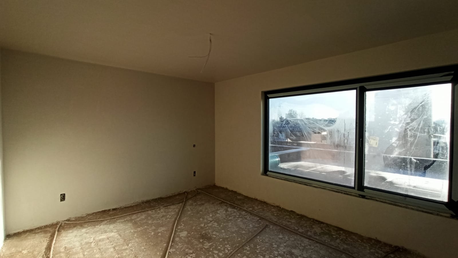 Pokój po nałożeniu jasnego tynku maszynowego, widoczne zabezpieczone okno oraz wystające kable elektryczne z sufitu.