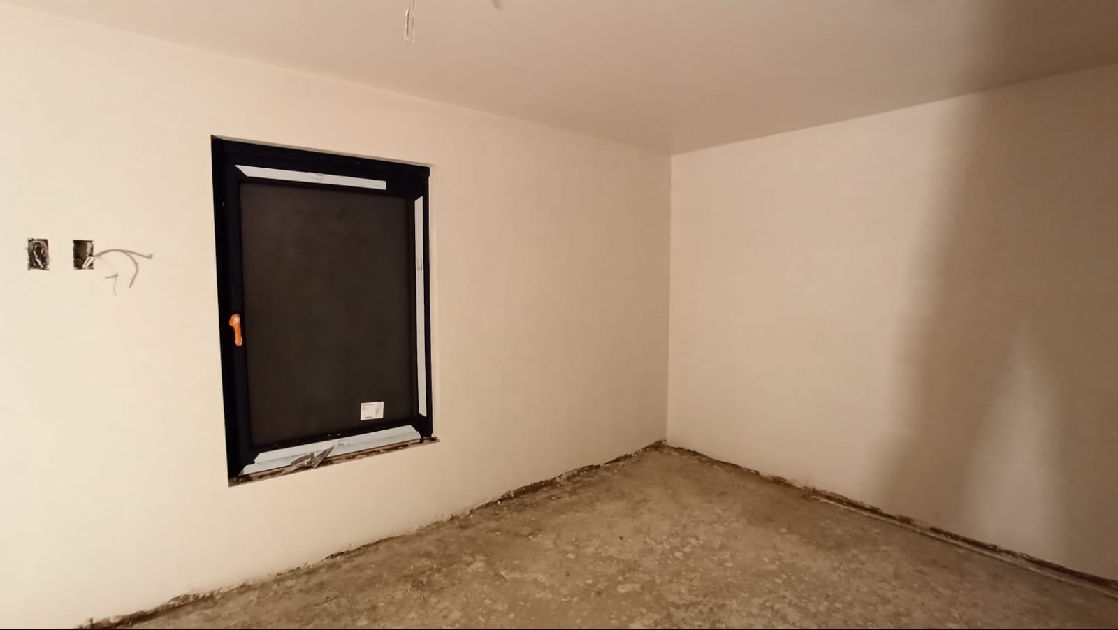 Pokój w trakcie tynkowania maszynowego: gładkie, jasne ściany, czarna rama okienna, widoczne puszki elektryczne z wystającymi przewodami oraz surowa, niezagruntowana podłoga.