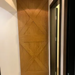 Jasne drewniane drzwi przesuwne w stylu rustykalnym z krzyżowym wzorem, zamontowane w wąskim korytarzu z szarą podłogą i widocznym fragmentem ościeżnicy drugich drzwi.