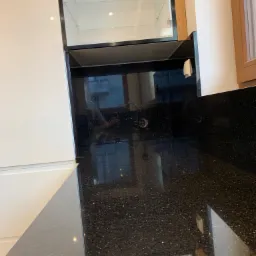 Fragment nowoczesnej kuchni z czarnym, błyszczącym blatem z drobnymi, srebrnymi inkluzjami, białymi szafkami i przeszkloną witryną górną.