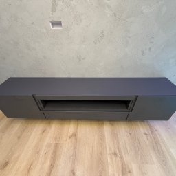 MSMeble (ZAKS) - Nowoczesna szafka RTV w kolorze grafitowym, zawieszona na ścianie o surowym wykończeniu, z jasną podłogą. Minimalistyczny design, brak uchwytów.