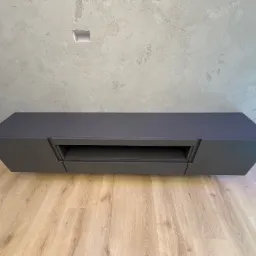 Nowoczesna szafka RTV w kolorze grafitowym, zawieszona na ścianie o surowym wykończeniu, z jasną podłogą. Minimalistyczny design, brak uchwytów.