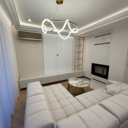 MSMeble (ZAKS) - Jasny salon z narożnikiem, stolikiem kawowym i zabudową ściany z kominkiem. Nowoczesna lampa sufitowa, minimalistyczny design.