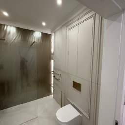 MSMeble (ZAKS) - Elegancka łazienka w odcieniach beżu i złota: kabina prysznicowa ze złotą armaturą, podwieszana toaleta i dekoracyjne panele ścienne z ramami.