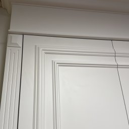 MSMeble (ZAKS) - Detal górnej części białej szafy z frezowanymi drzwiami i ozdobnym gzymsem, widok z dołu, ukazujący precyzję wykonania.