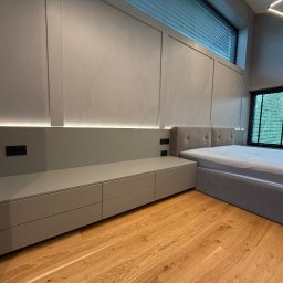 MSMeble (ZAKS) - Nowoczesna sypialnia z szarą zabudową meblową: szafki pod oknem, wezgłowie łóżka. Drewniana podłoga, stonowana kolorystyka, minimalistyczne oświetlenie LED.