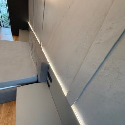 MSMeble (ZAKS) - Nowoczesna sypialnia: szara tapicerowana rama łóżka z wbudowanymi gniazdkami, minimalistyczna szafka nocna i dekoracyjna ściana z podświetleniem LED.