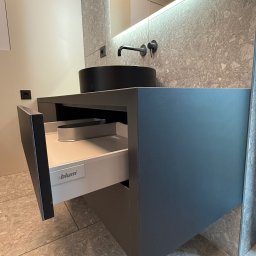 MSMeble (ZAKS) - Szara szafka łazienkowa z wysuniętą szufladą Blum, czarna umywalka nablatowa i podświetlane lustro na tle ściany z szarego kamienia.