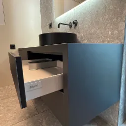 Szara szafka łazienkowa z wysuniętą szufladą Blum, czarna umywalka nablatowa i podświetlane lustro na tle ściany z szarego kamienia.