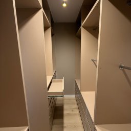 MSMeble (ZAKS) - Beżowa garderoba na wymiar z wysuwaną szufladą, oświetleniem punktowym i drążkami na ubrania. Widok z perspektywy wejścia, ukazujący symetryczny układ półek i szuflad.