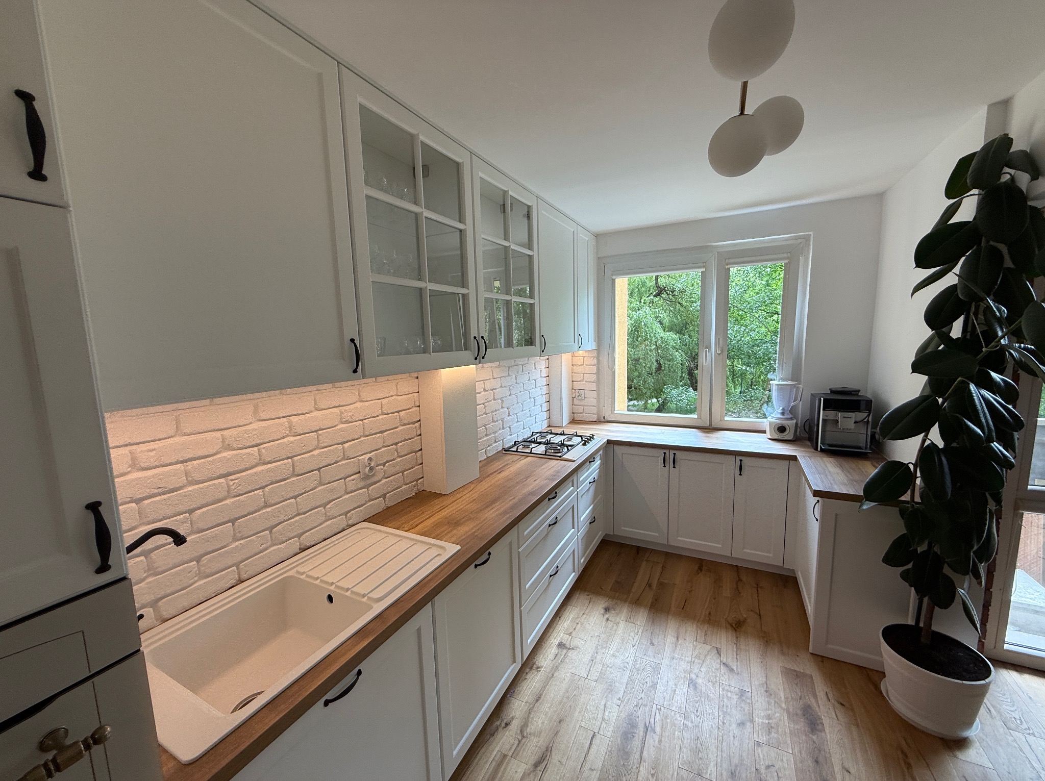 Jasna kuchnia w stylu rustykalnym z drewnianym blatem, białymi szafkami z ciemnymi uchwytami i ceglaną ścianą. Widok na ogród przez okno.