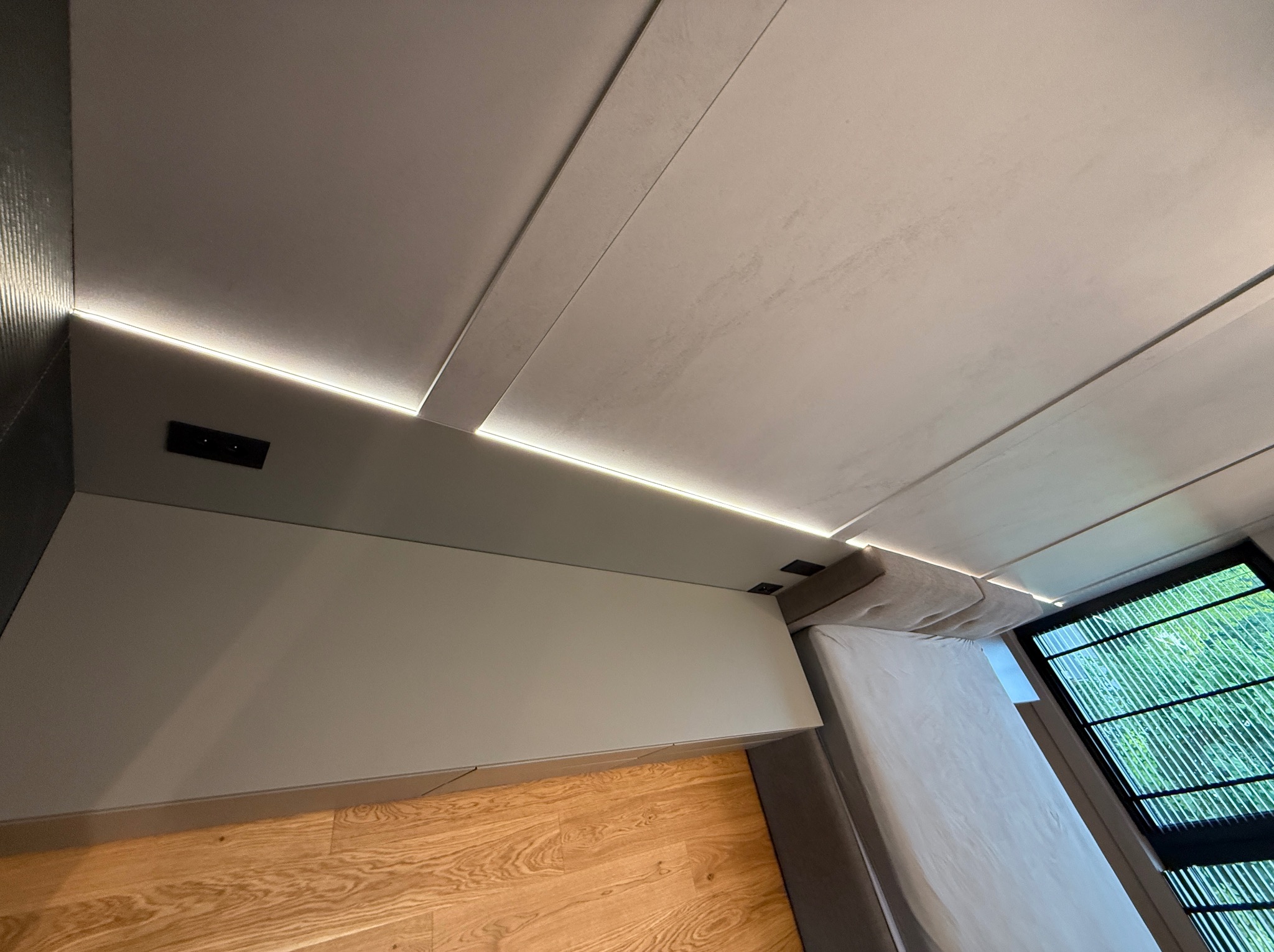 Nowoczesna sypialnia: drewniana podłoga, szara ściana z listwą LED, łóżko i okno z widokiem na zieleń. Minimalistyczny design.