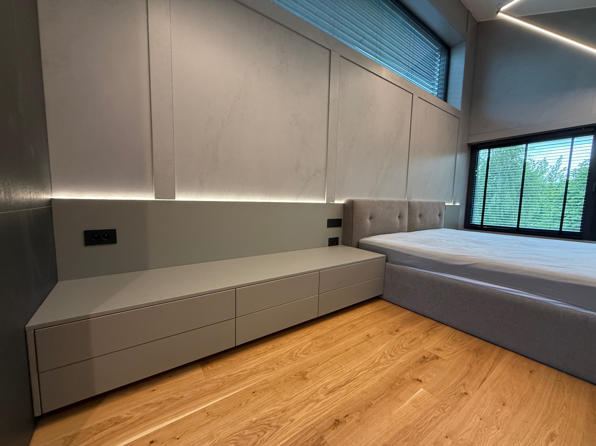 Nowoczesna sypialnia z szarą zabudową meblową: szafki pod oknem, wezgłowie łóżka. Drewniana podłoga, stonowana kolorystyka, minimalistyczne oświetlenie LED.