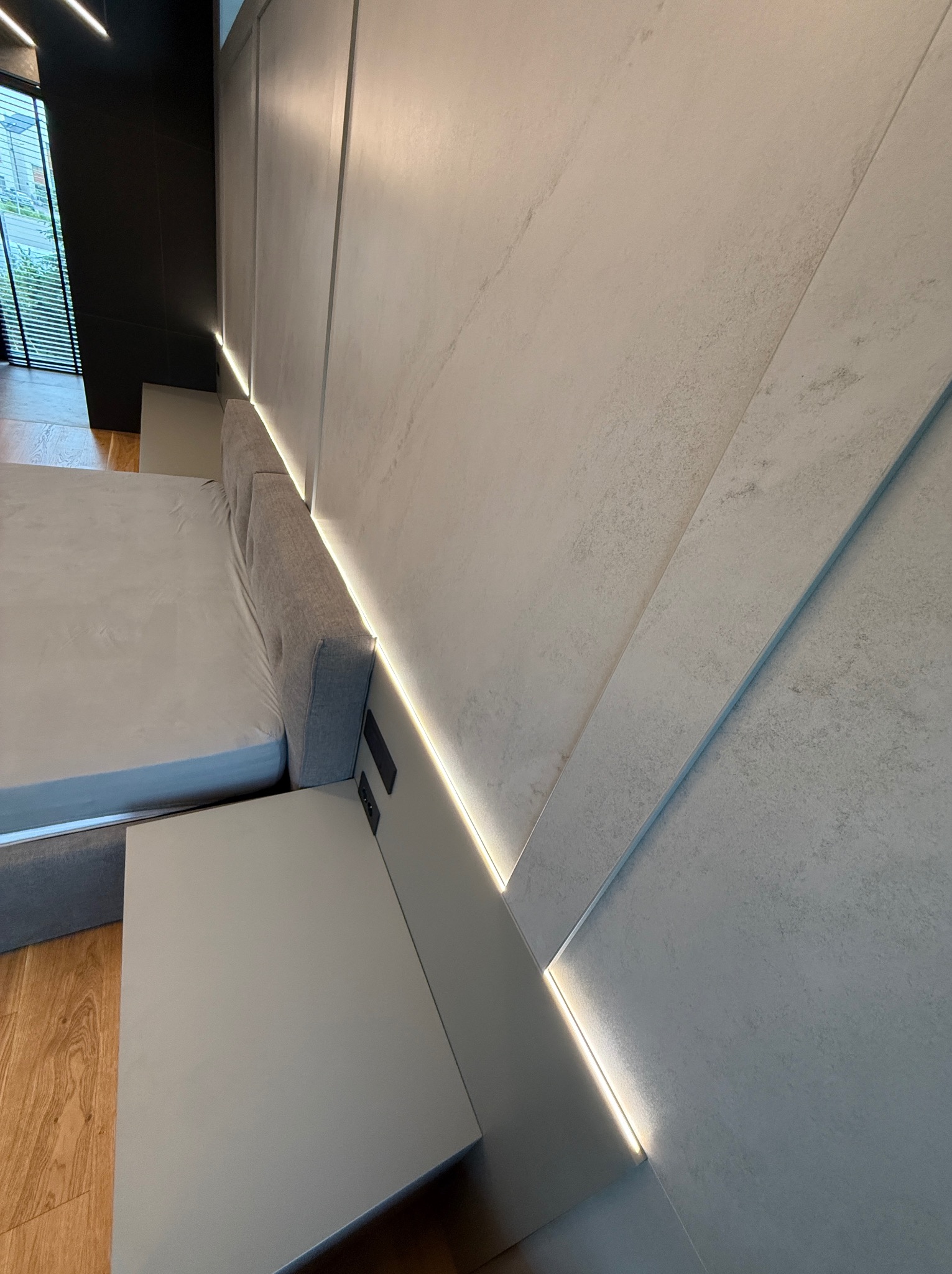 Nowoczesna sypialnia: szara tapicerowana rama łóżka z wbudowanymi gniazdkami, minimalistyczna szafka nocna i dekoracyjna ściana z podświetleniem LED.