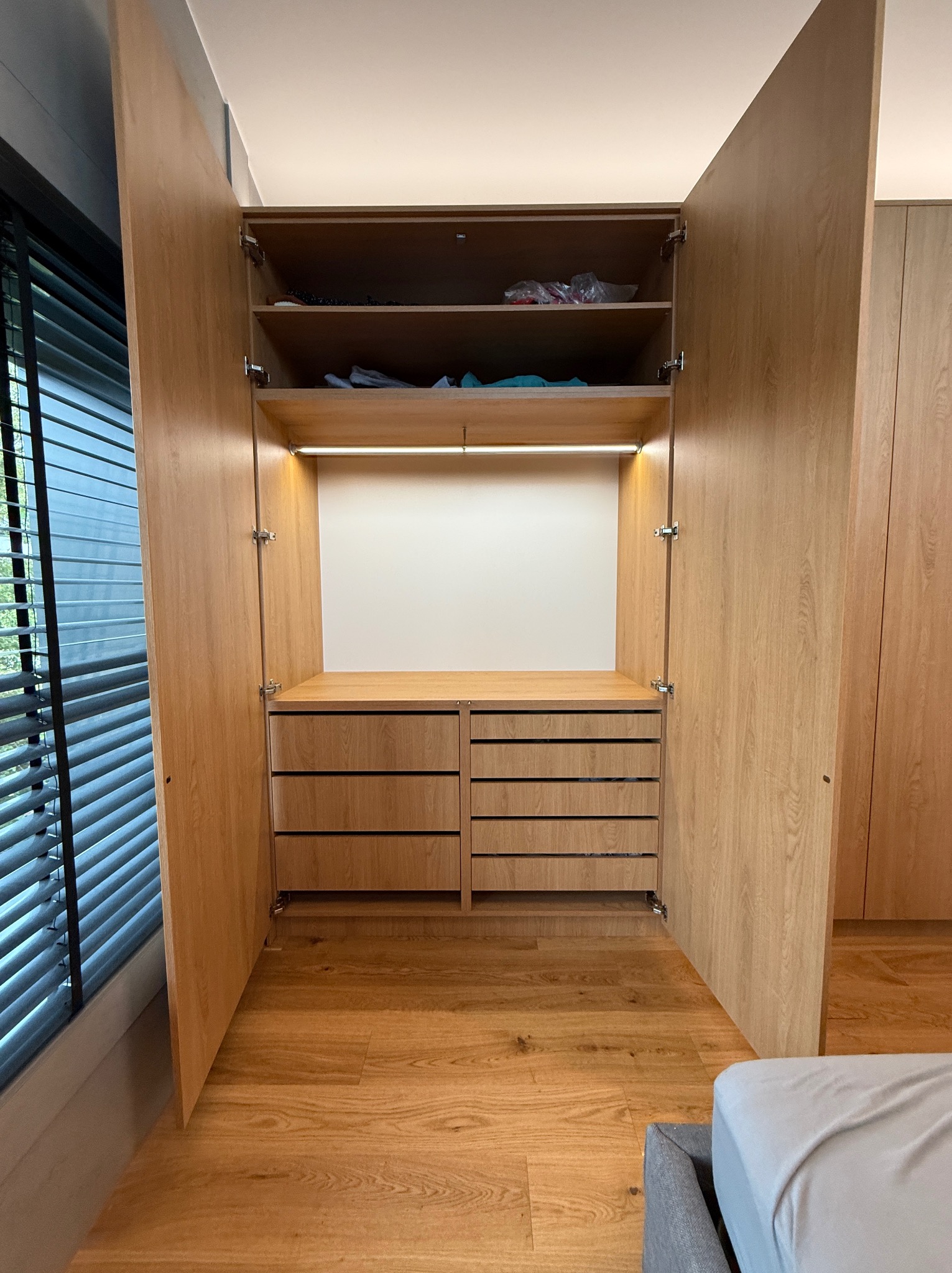 Otwarta szafa wnękowa z jasnego drewna, z półkami i szufladami, oświetlona od wewnątrz. Widoczna podłoga z paneli i fragment okna z żaluzjami.