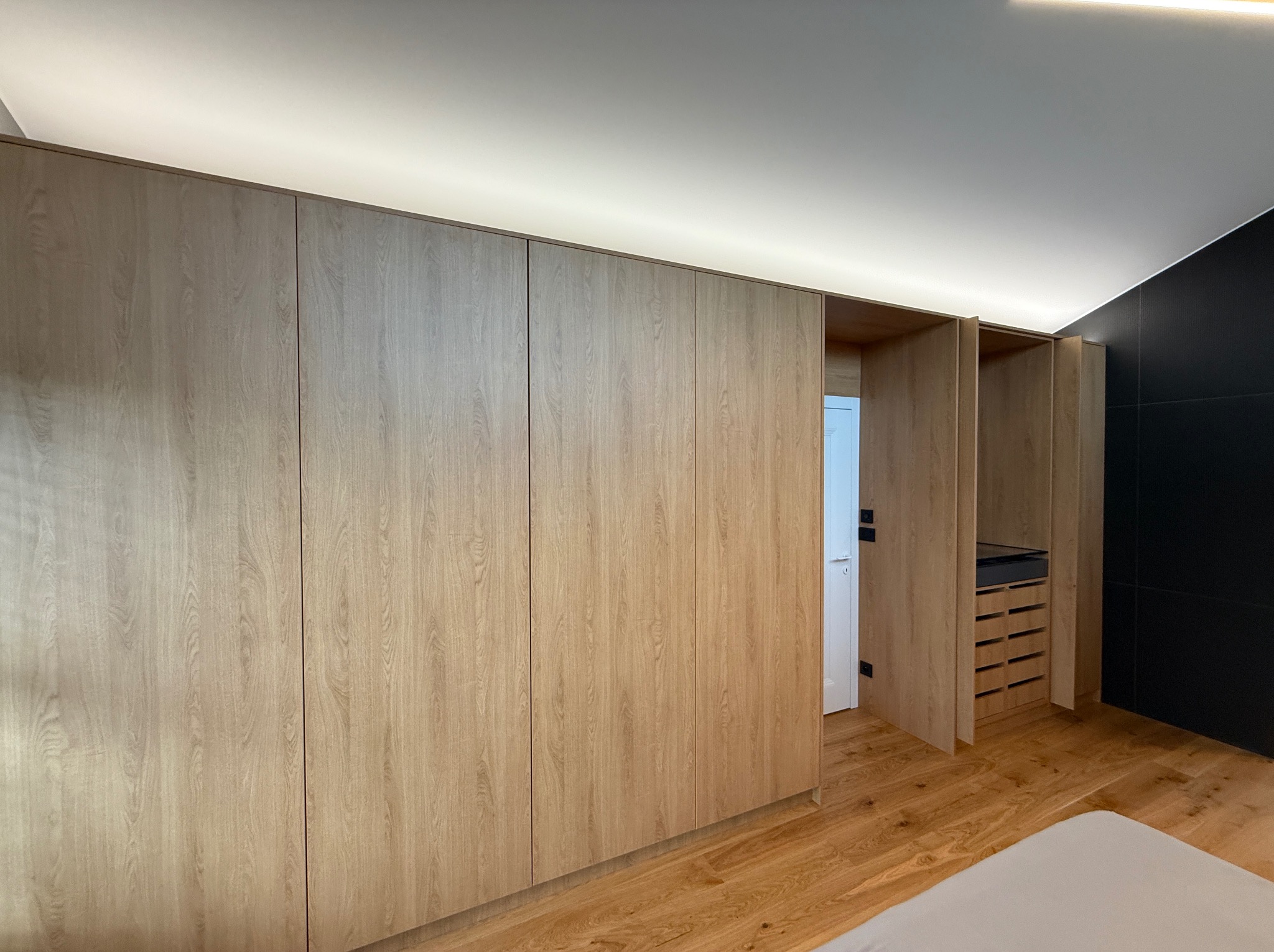 Jasna szafa wnękowa z drewnianymi frontami, oświetleniem LED u góry i częściowo otwartymi drzwiami, ukazująca wnętrze z szufladami. Minimalistyczny design.