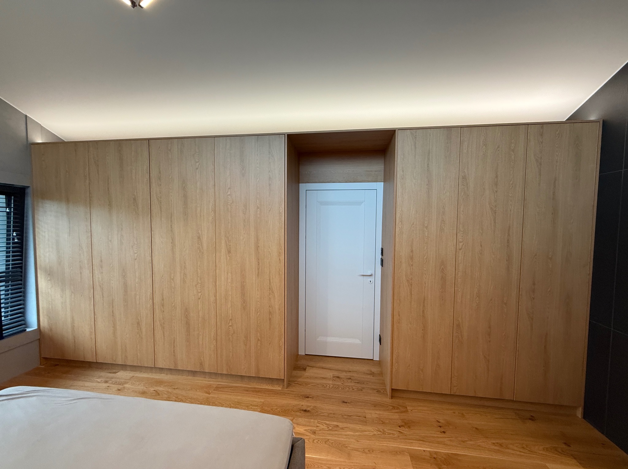 Jasna, drewniana szafa na wymiar z wnęką na białe drzwi w sypialni. Widoczne fragmenty łóżka i podłogi z paneli. Oświetlenie LED podszafkowe.