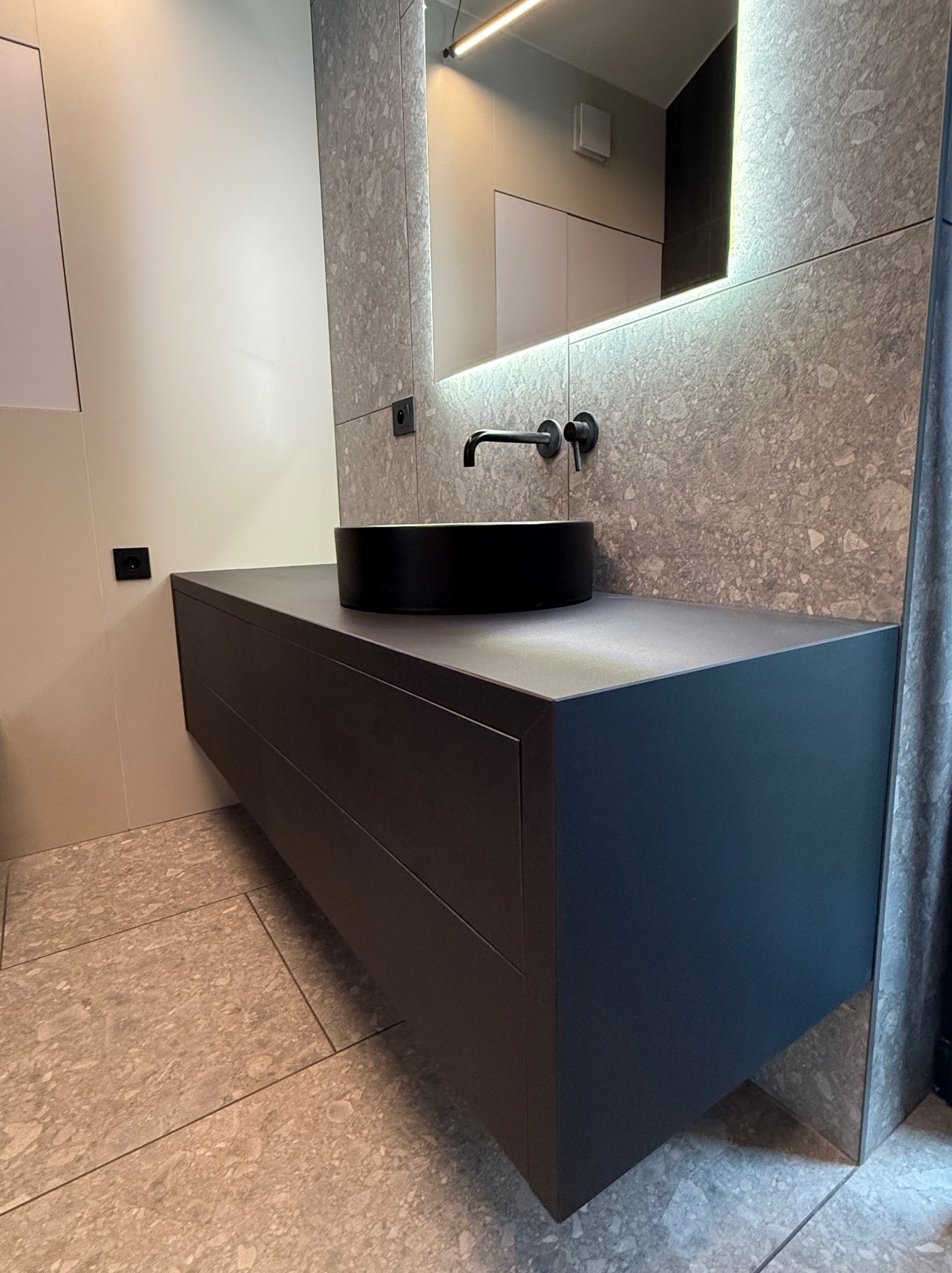 Nowoczesna łazienka: ciemna szafka z umywalką typu misa, czarna armatura na tle ściany z płytek imitujących kamień i lustra z podświetleniem LED.