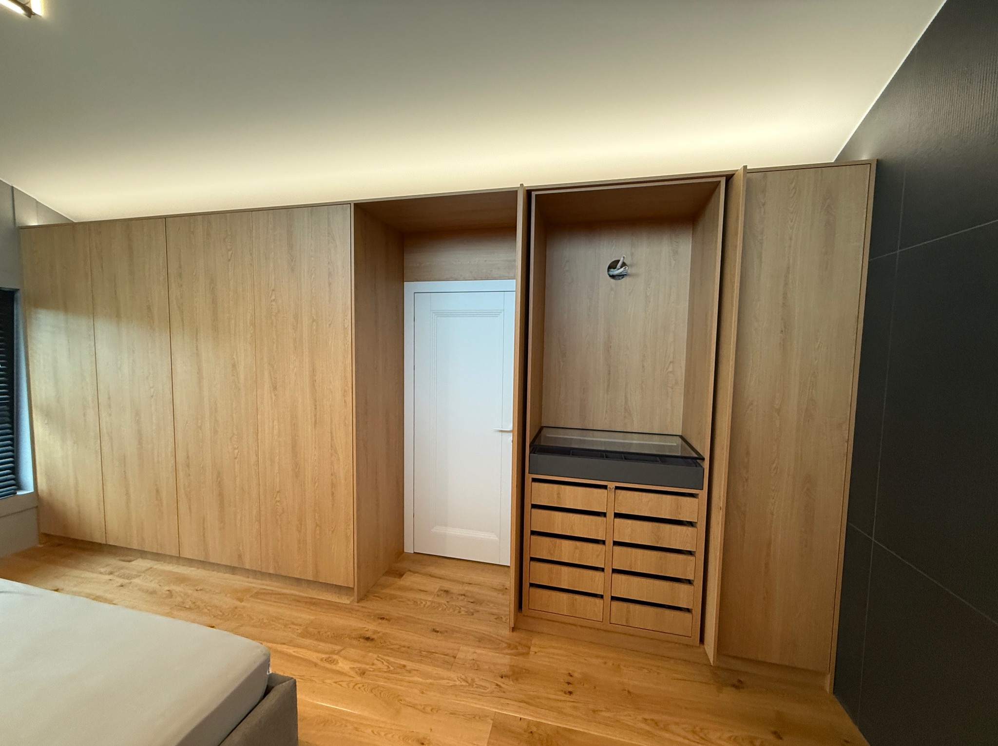 Jasna, drewniana szafa wnękowa z szufladami i otwartą przestrzenią na drzwi w tle. Podłoga z paneli w odcieniach drewna. Ciemna ściana z prawej strony.