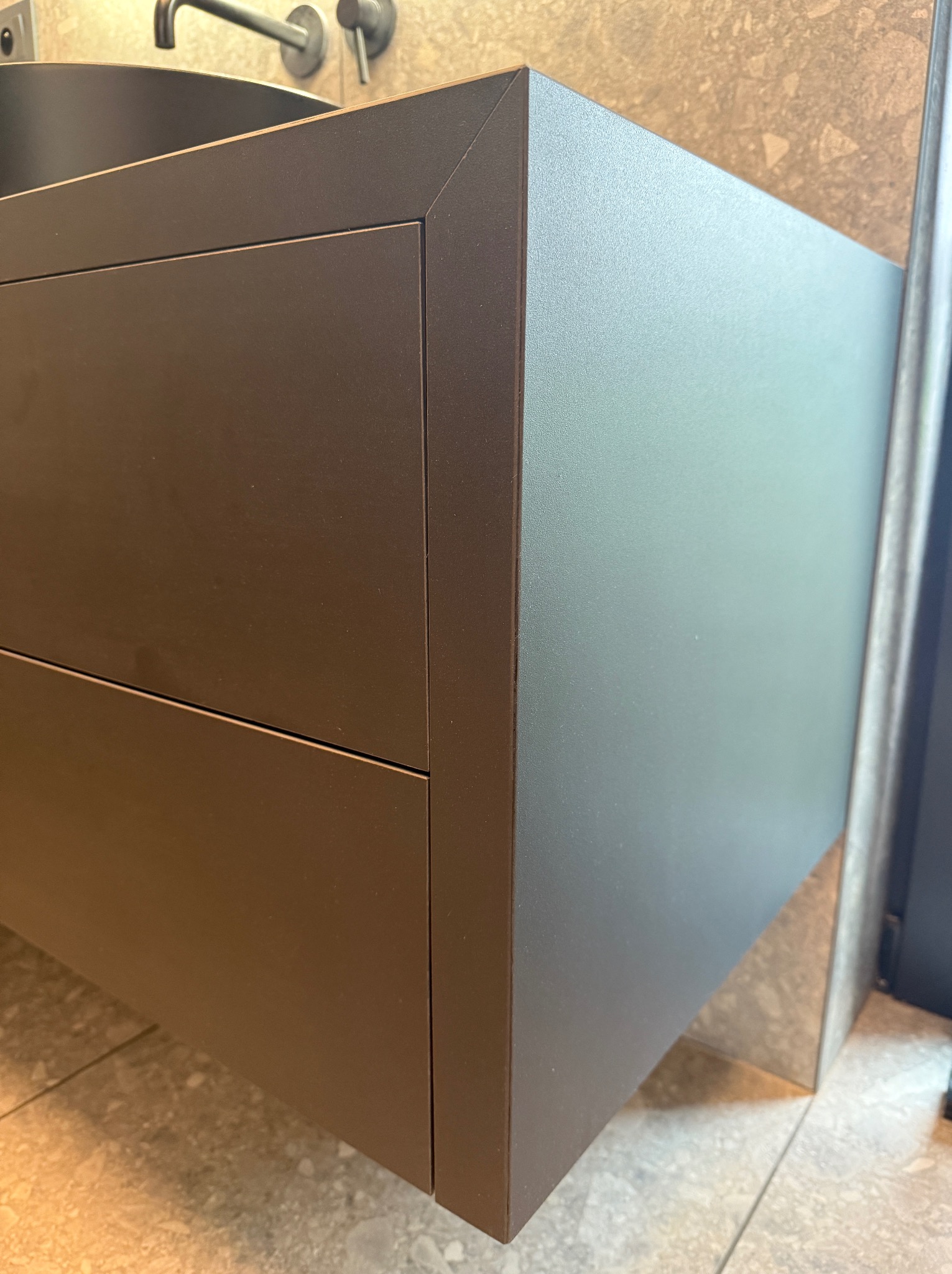 Nowoczesna, grafitowa szafka łazienkowa z dwiema szufladami, zintegrowana z umywalką i baterią, na tle beżowych płytek. Minimalistyczny design.