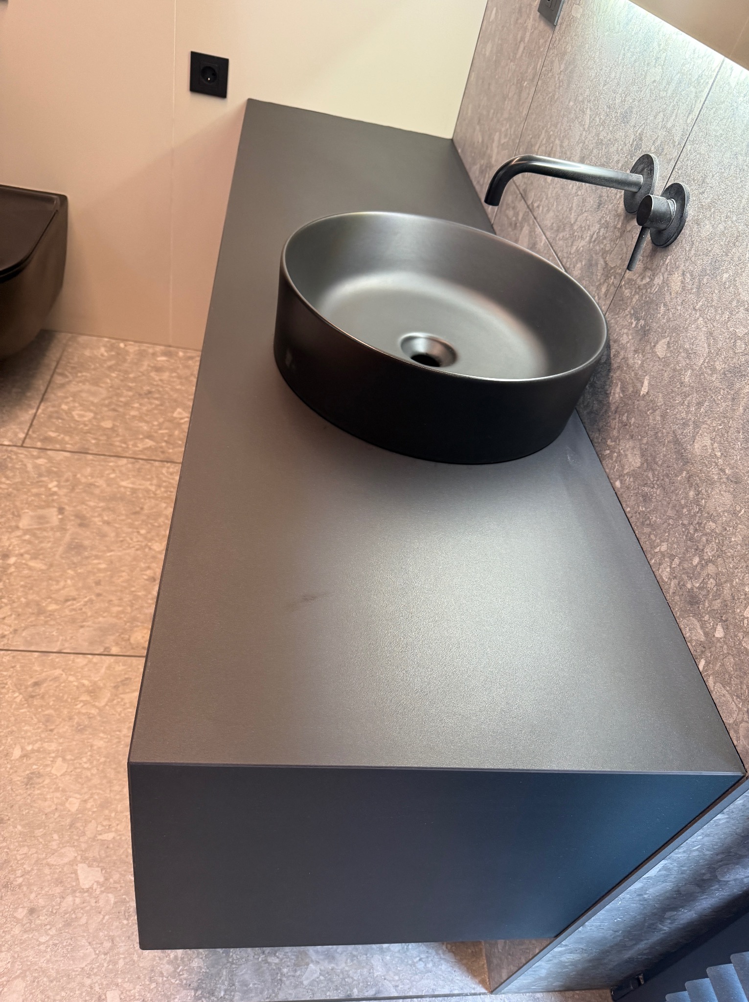 Szara, okrągła umywalka nablatowa na blacie w łazience, z nowoczesną armaturą i płytkami imitującymi kamień. Minimalistyczny design.