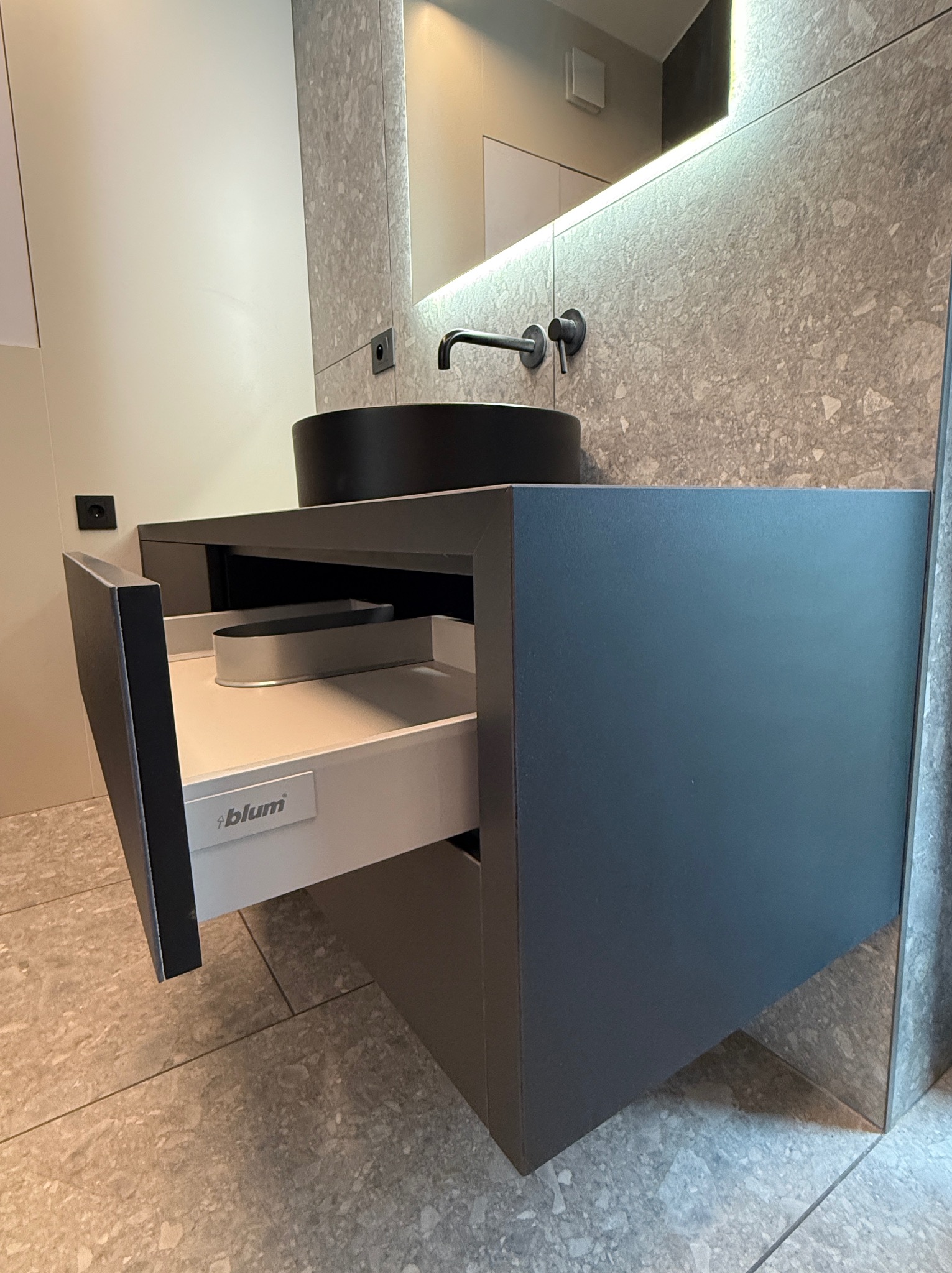 Szara szafka łazienkowa z wysuniętą szufladą Blum, czarna umywalka nablatowa i podświetlane lustro na tle ściany z szarego kamienia.