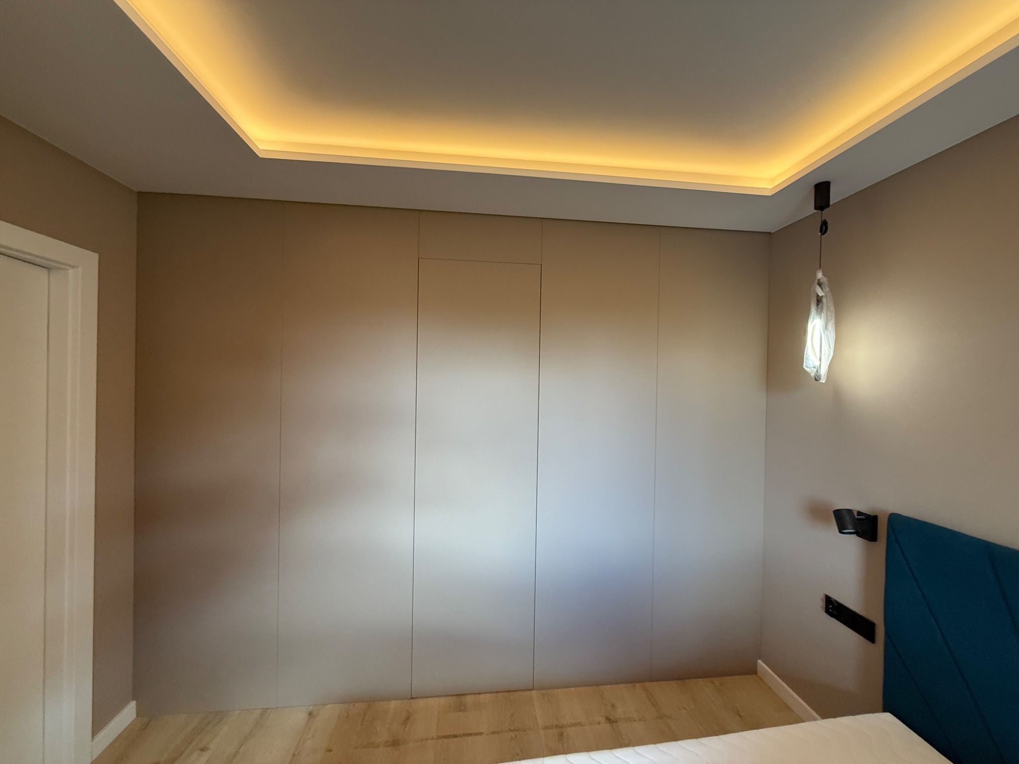 Beżowa szafa wnękowa z ukrytymi drzwiami, podświetlany sufit w sypialni z drewnianą podłogą. Minimalistyczny design z lampą wiszącą i wezgłowiem łóżka.