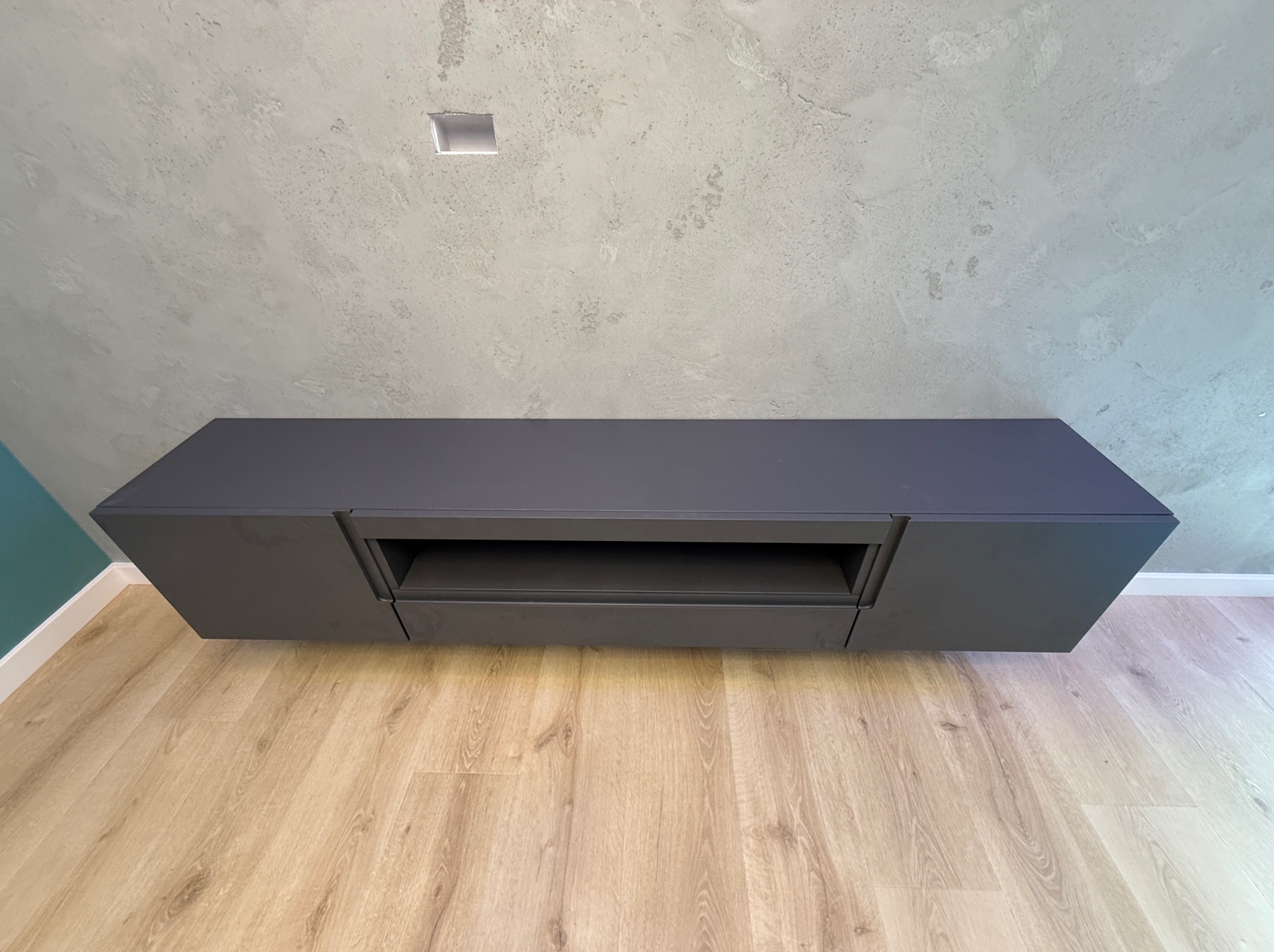 Grafitowa szafka RTV podwieszana na tle szarej ściany o betonowej fakturze i jasnej podłogi. Minimalistyczny design z widoczną półką. Element wystroju wnętrza.