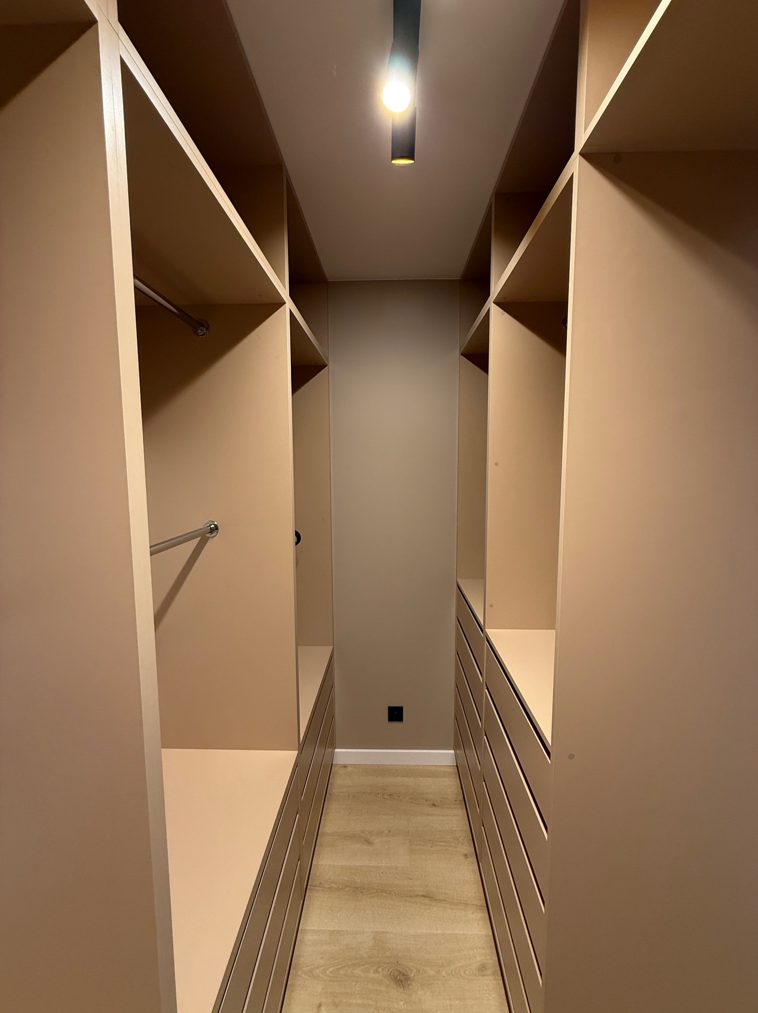 Beżowa garderoba na wymiar z półkami, drążkami i szufladami, oświetlona punktowo, widok z perspektywy wnętrza, na podłodze panele.