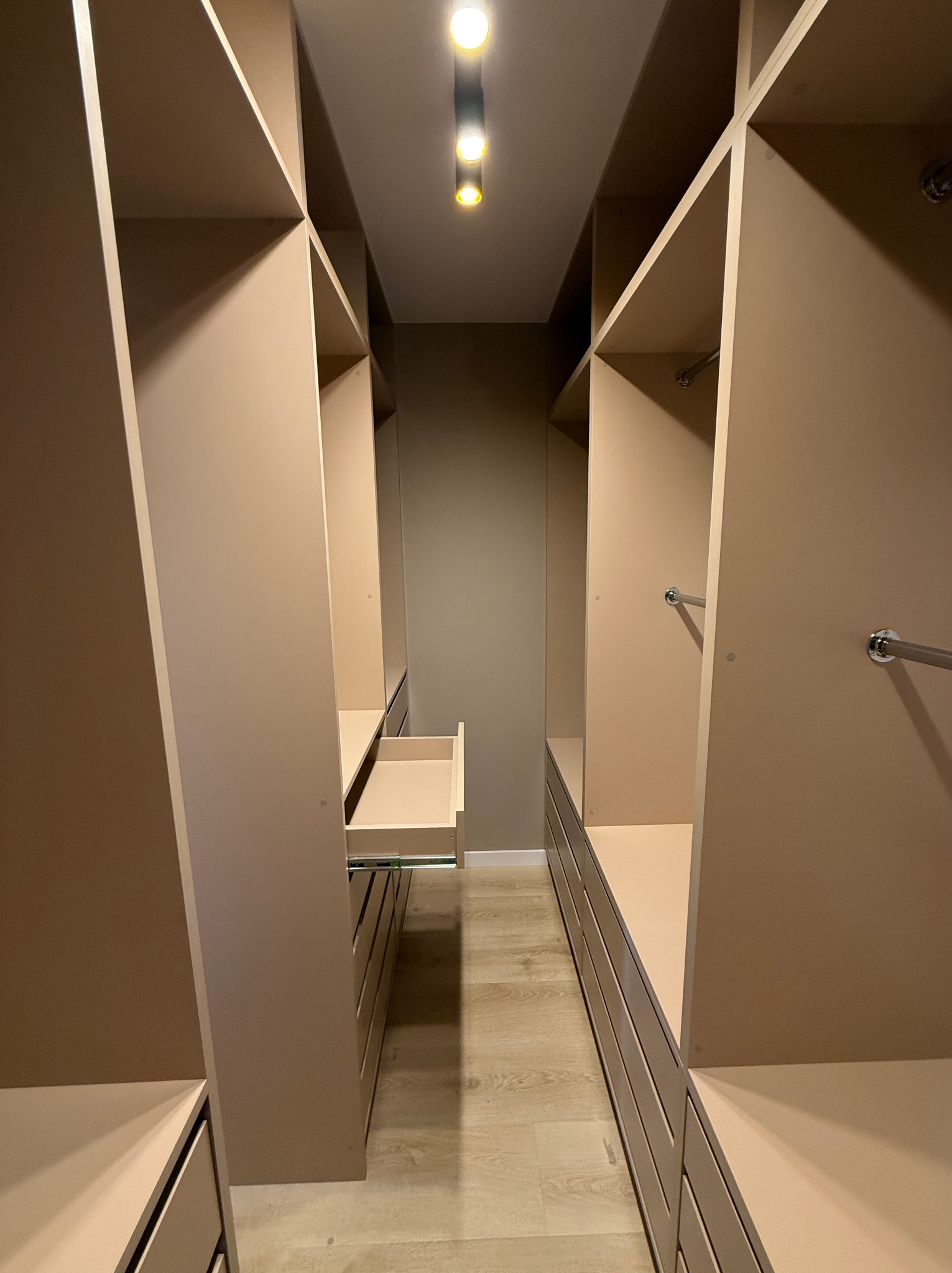 Beżowa garderoba na wymiar z wysuwaną szufladą, oświetleniem punktowym i drążkami na ubrania. Widok z perspektywy wejścia, ukazujący symetryczny układ półek i szuflad.