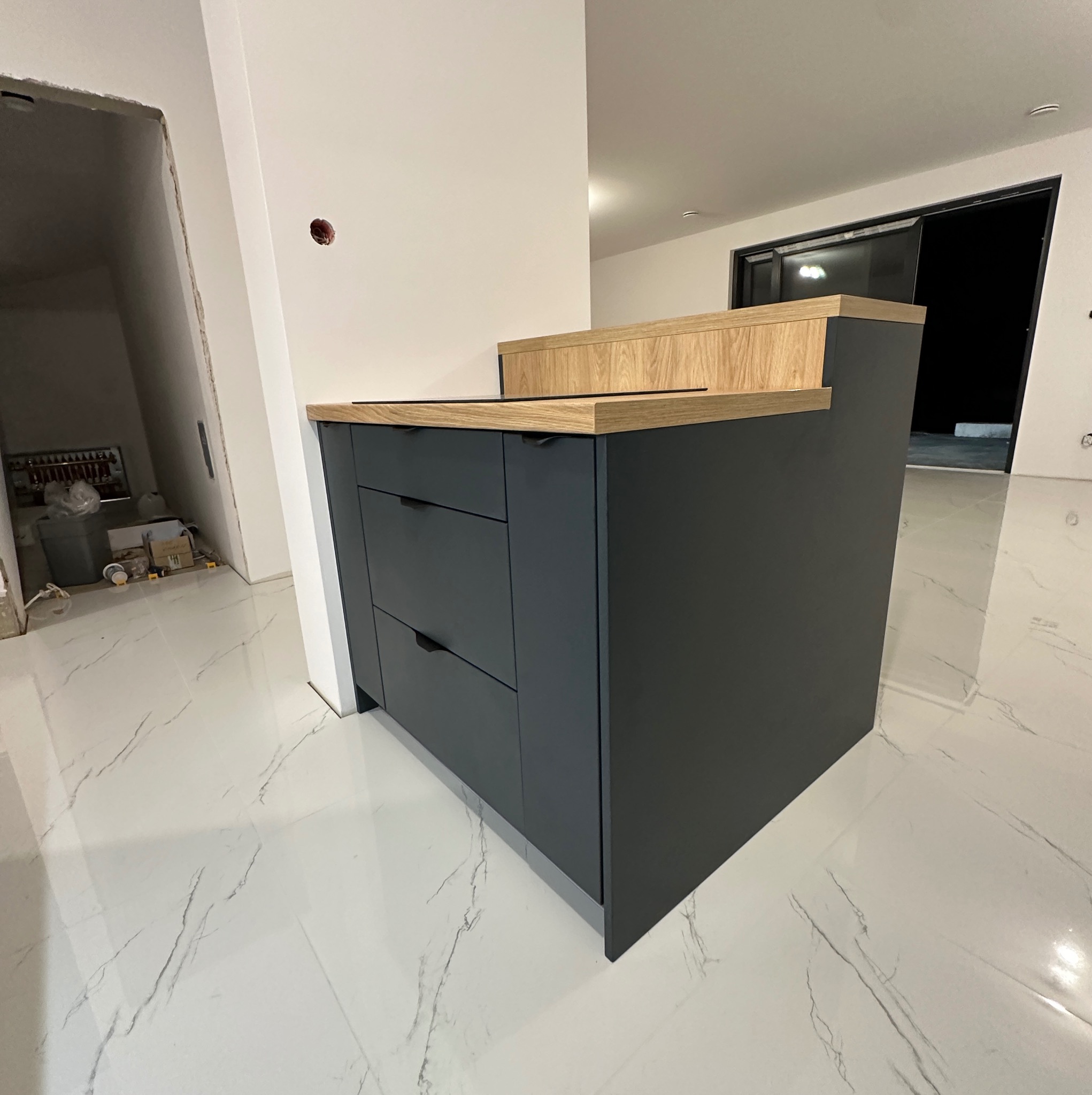 Nowoczesna, grafitowa szafka kuchenna z drewnianym blatem, widoczna na tle białej marmurowej podłogi w surowym wnętrzu.