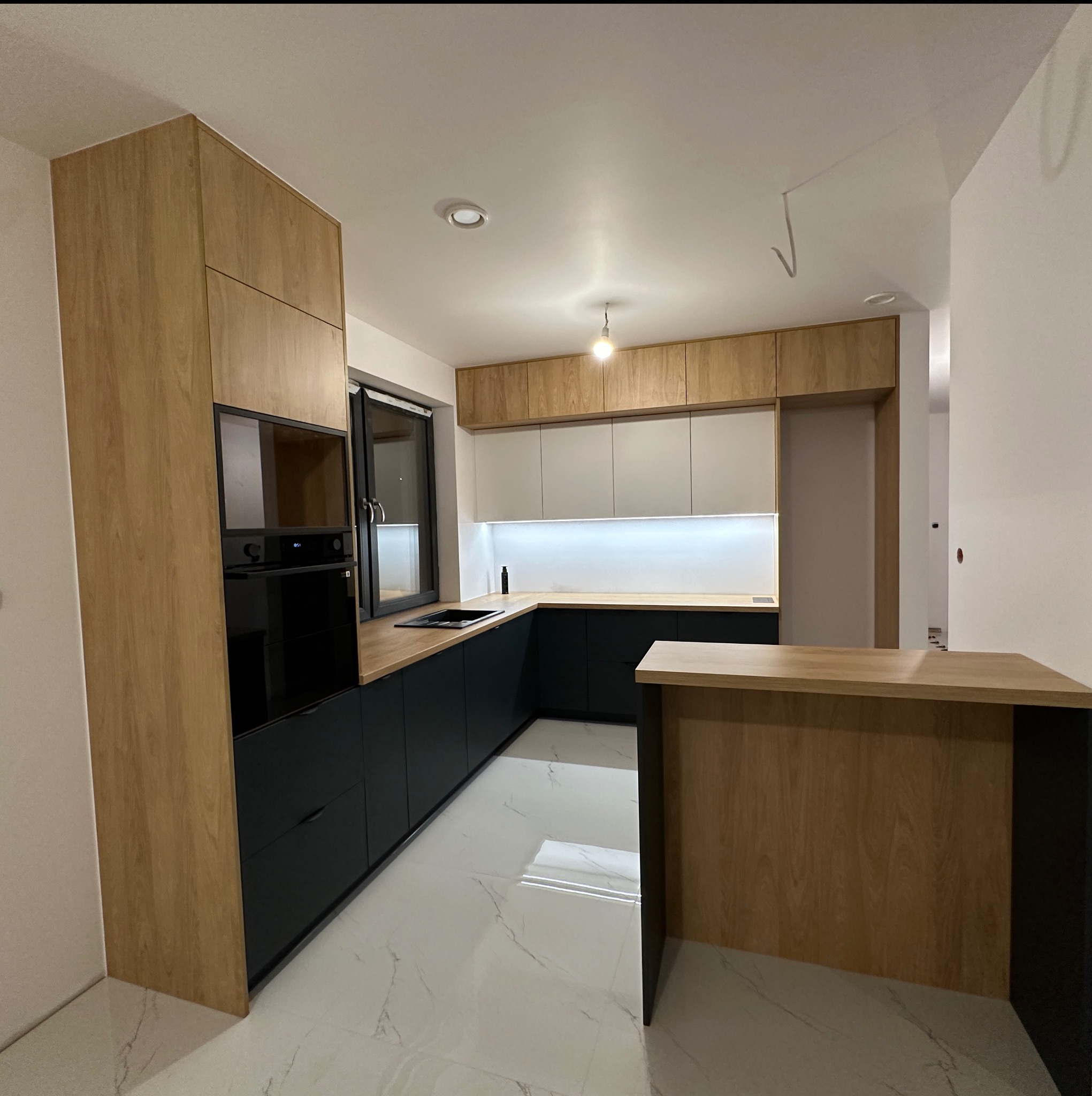 Nowoczesna kuchnia w Radomsku z drewnianymi frontami i czarnymi dolnymi szafkami. Uwagę przyciąga barek z blatem w kolorze drewna i marmurowa podłoga.