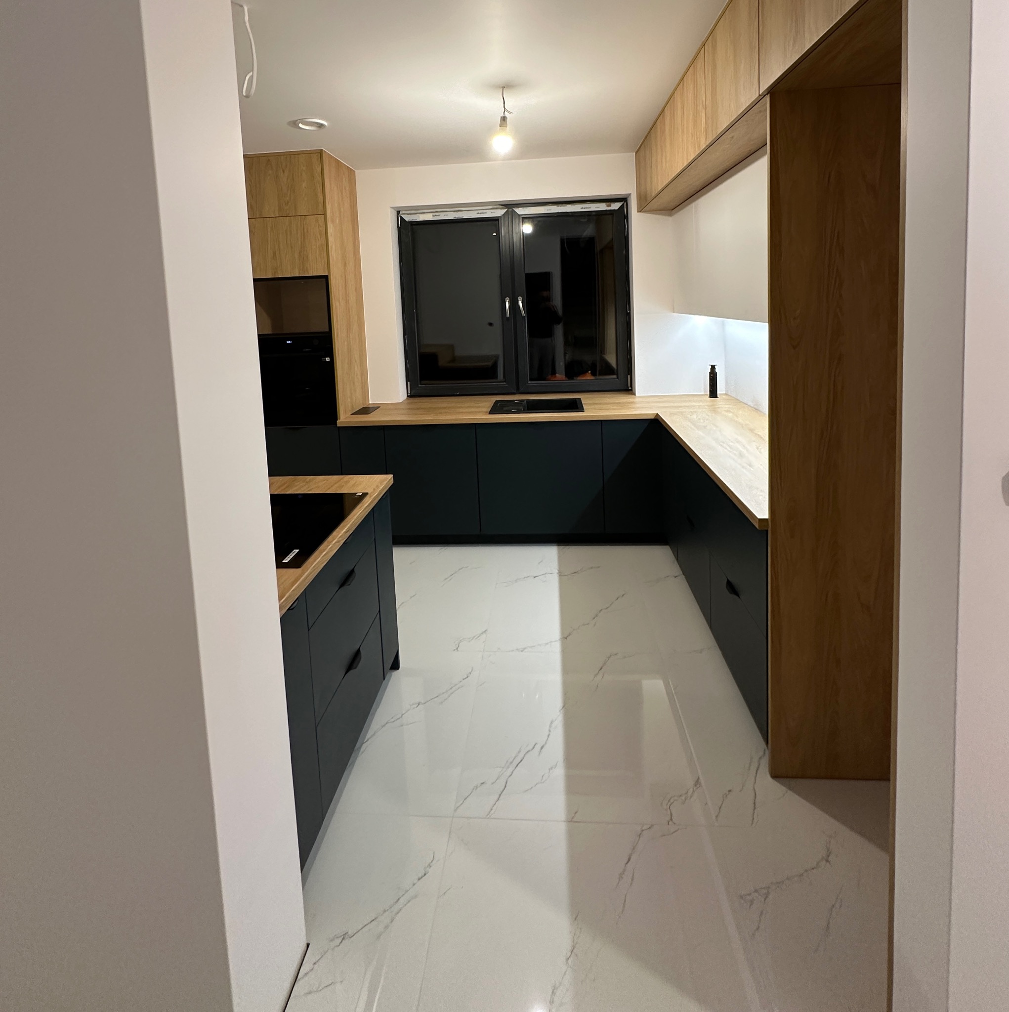 Nowoczesna kuchnia z ciemnymi szafkami i drewnianymi blatami, widok z perspektywy wejścia. Białe marmurowe płytki podłogowe, czarne okno.