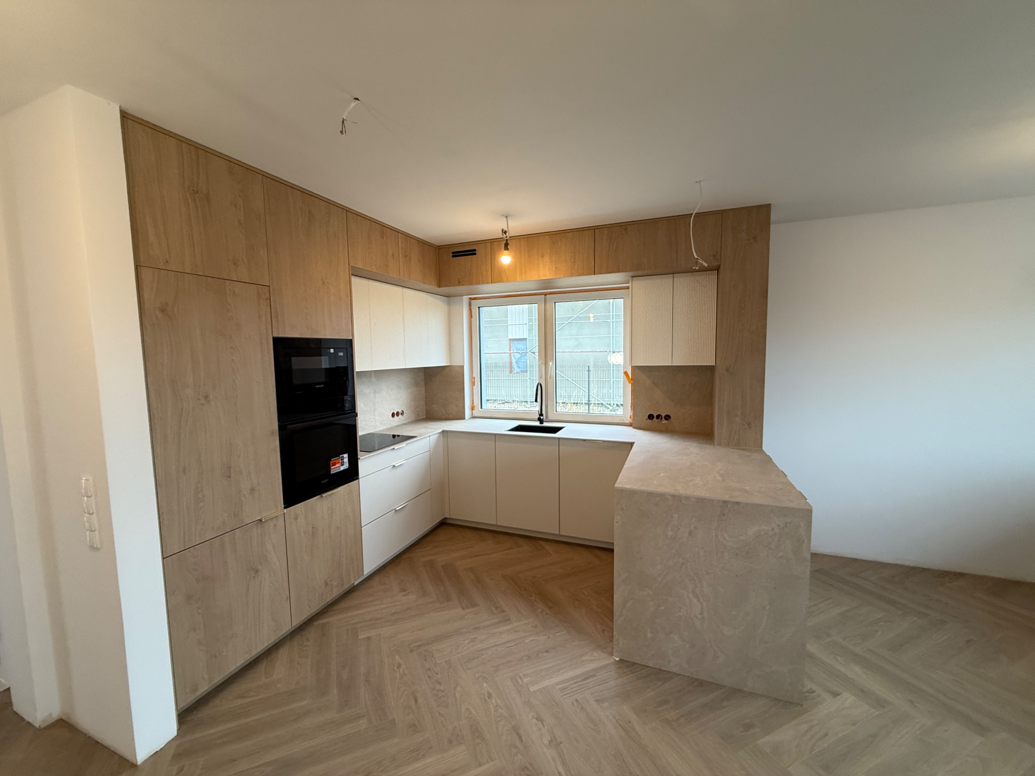 Nowoczesna kuchnia z drewnianymi i białymi frontami, czarnym piekarnikiem i blatem w odcieniach beżu. Parkiet ułożony w jodełkę. Widoczne gniazdka elektryczne.