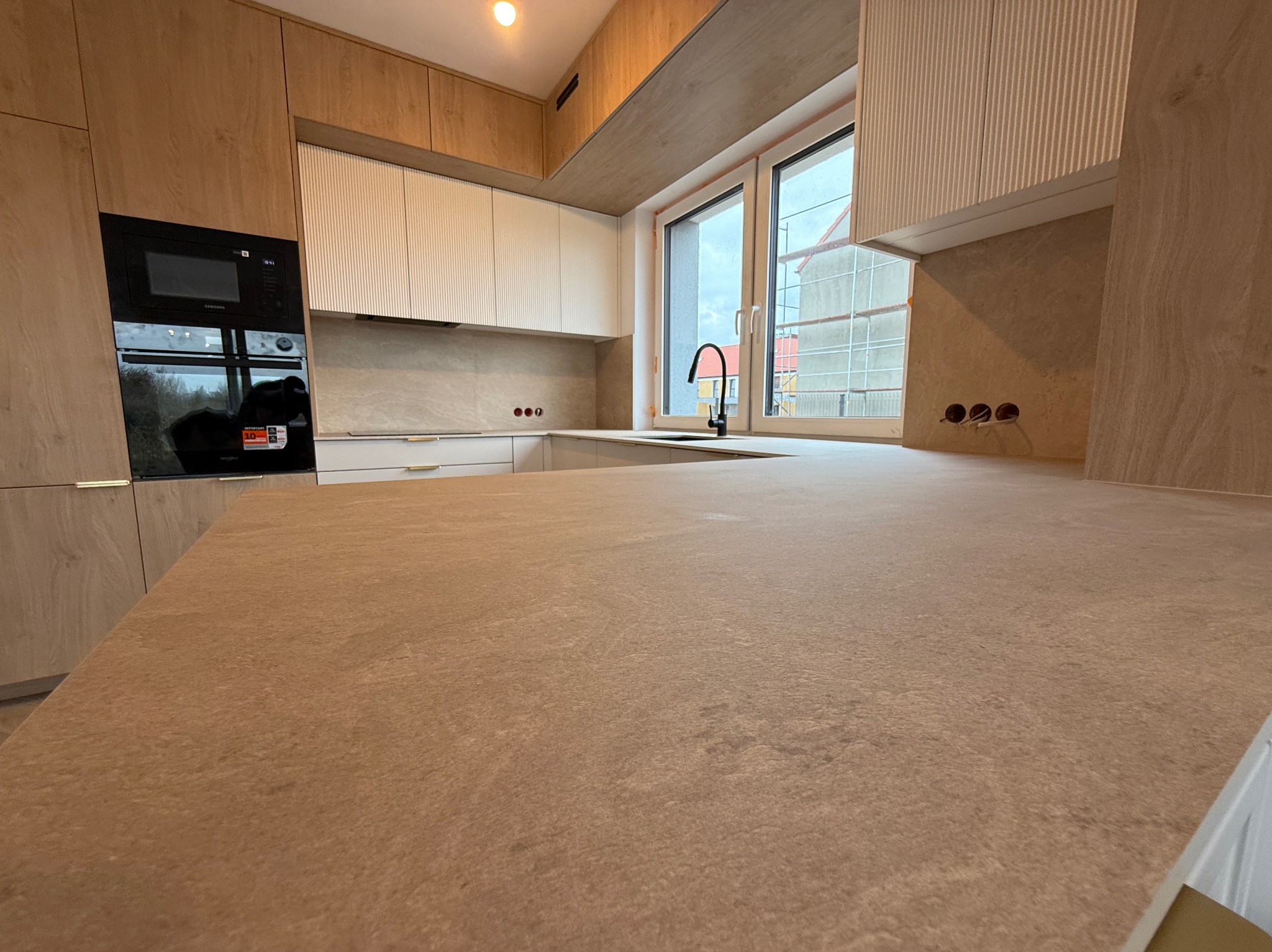 Jasna kuchnia na wymiar z blatem w odcieniach beżu. Uwagę przykuwają ryflowane fronty szafek wiszących i nowoczesna bateria kuchenna.