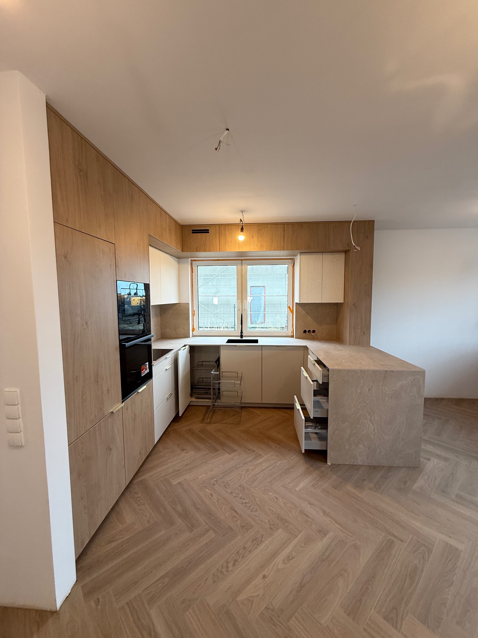 Jasna kuchnia z drewnianymi frontami i wyspą na parkiecie w jodełkę, widok na okno z czarną armaturą, nowoczesny design, oświetlenie sufitowe.