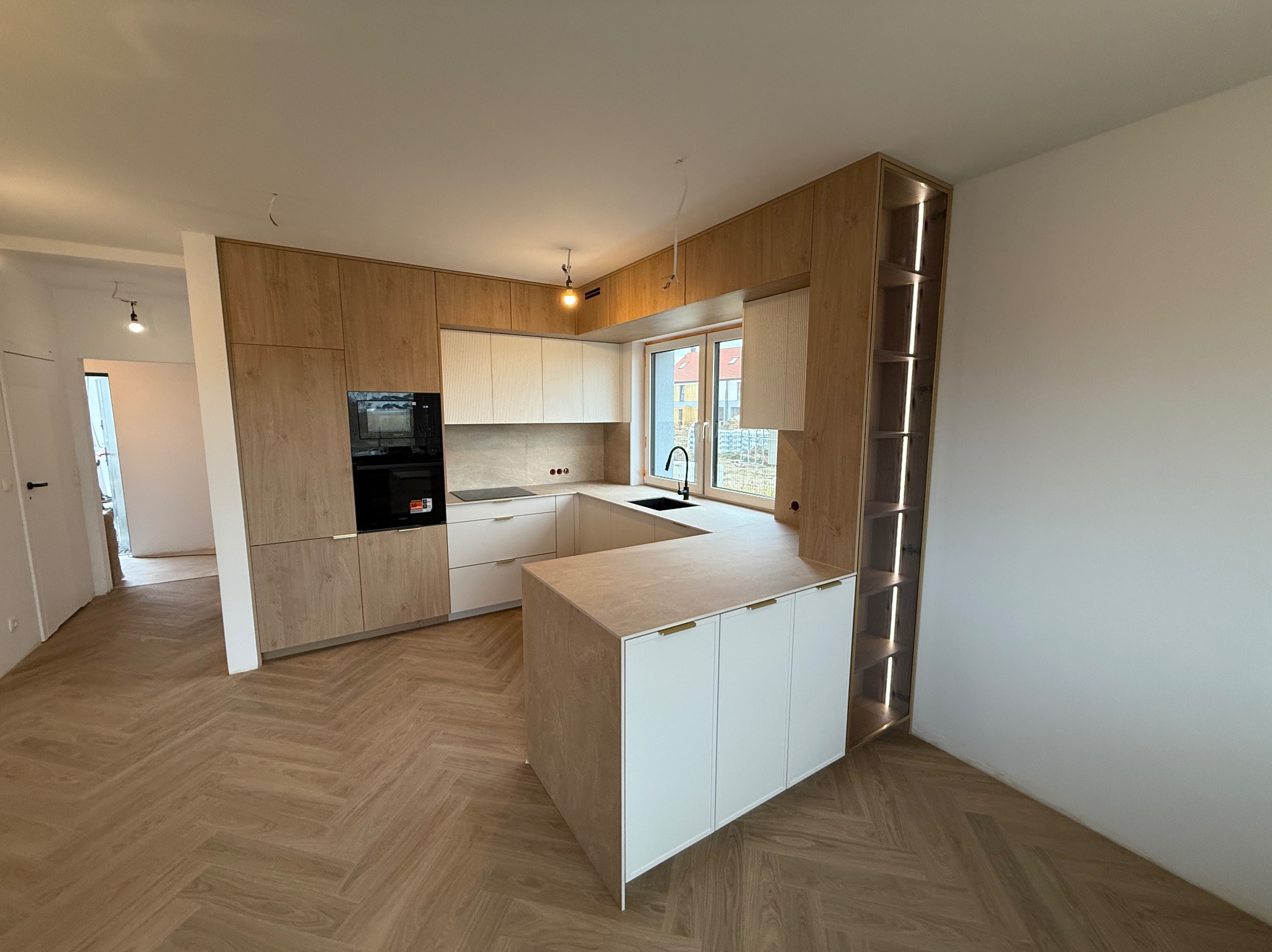 Nowoczesna kuchnia z wyspą, drewniane fronty szafek górnych i dolnych, białe szafki na wyspie, oświetlenie LED w regale, parkiet w jodełkę, czarny zlew.