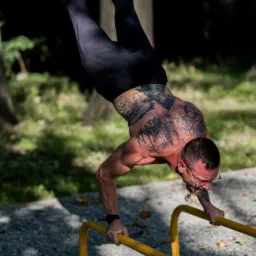 Mężczyzna z tatuażami na plecach i ramionach, w okularach, wykonuje stójkę na rękach na żółtych poręczach w parku, w tle drzewa i trawa.