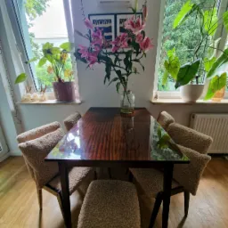 Jadalnia z drewnianym stołem i krzesłami obitymi beżową tkaniną typu baranek, na stole wazon z różowymi liliami, w tle okna z widokiem na zieleń i rośliny doniczkowe na parapetach.