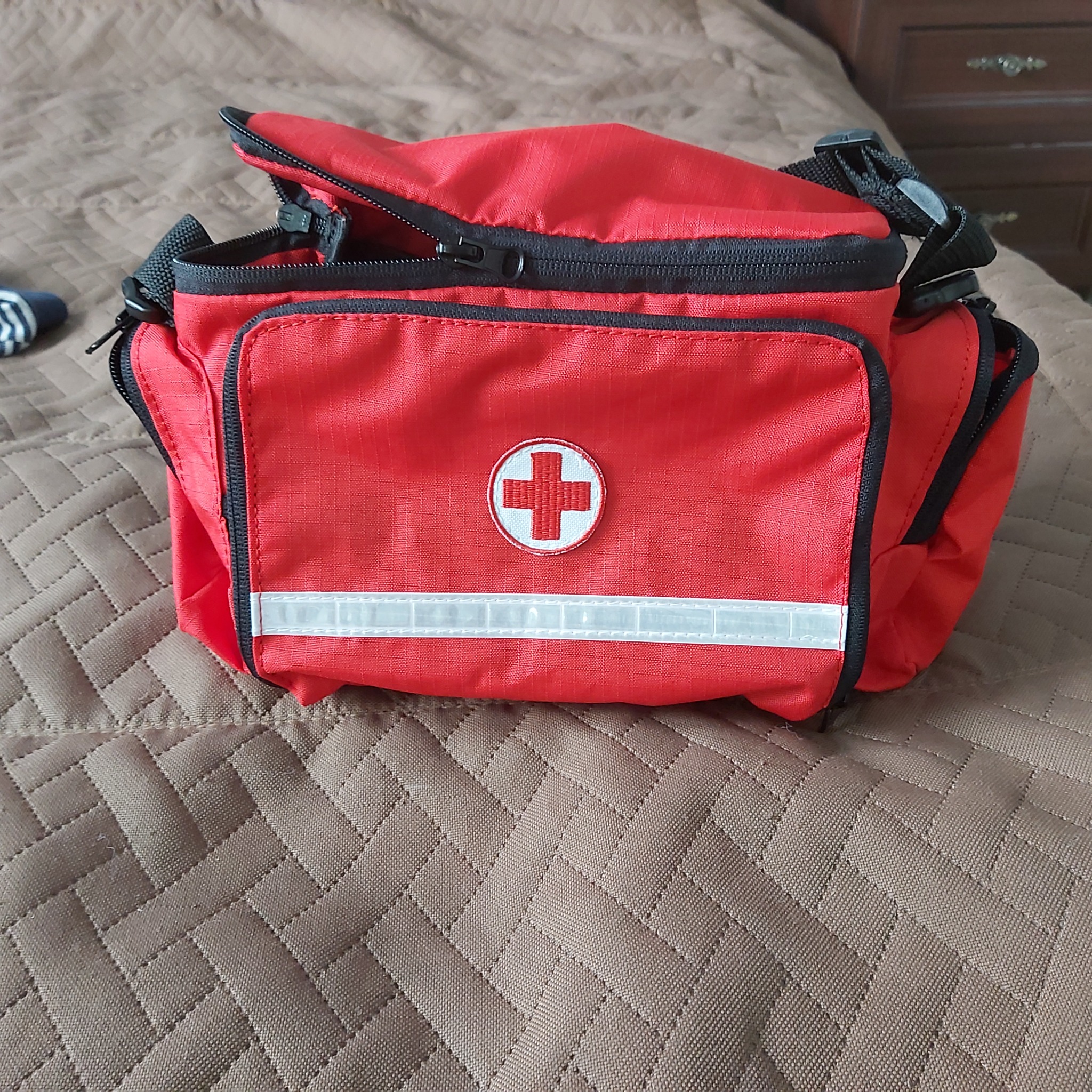 Czerwona torba medyczna z białym krzyżem i odblaskowym paskiem, leżąca na brązowej, pikowanej narzucie.