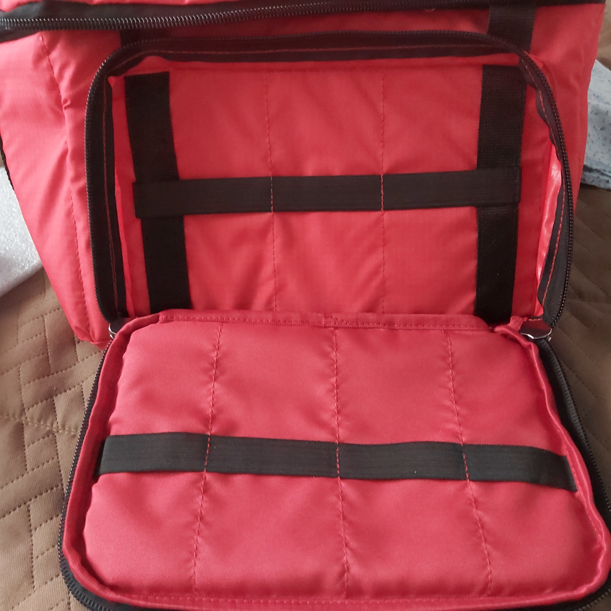 Otwarta czerwona torba z czarnymi elastycznymi paskami wewnątrz, widoczne szwy i zamek błyskawiczny, leżąca na brązowej pikowanej powierzchni.