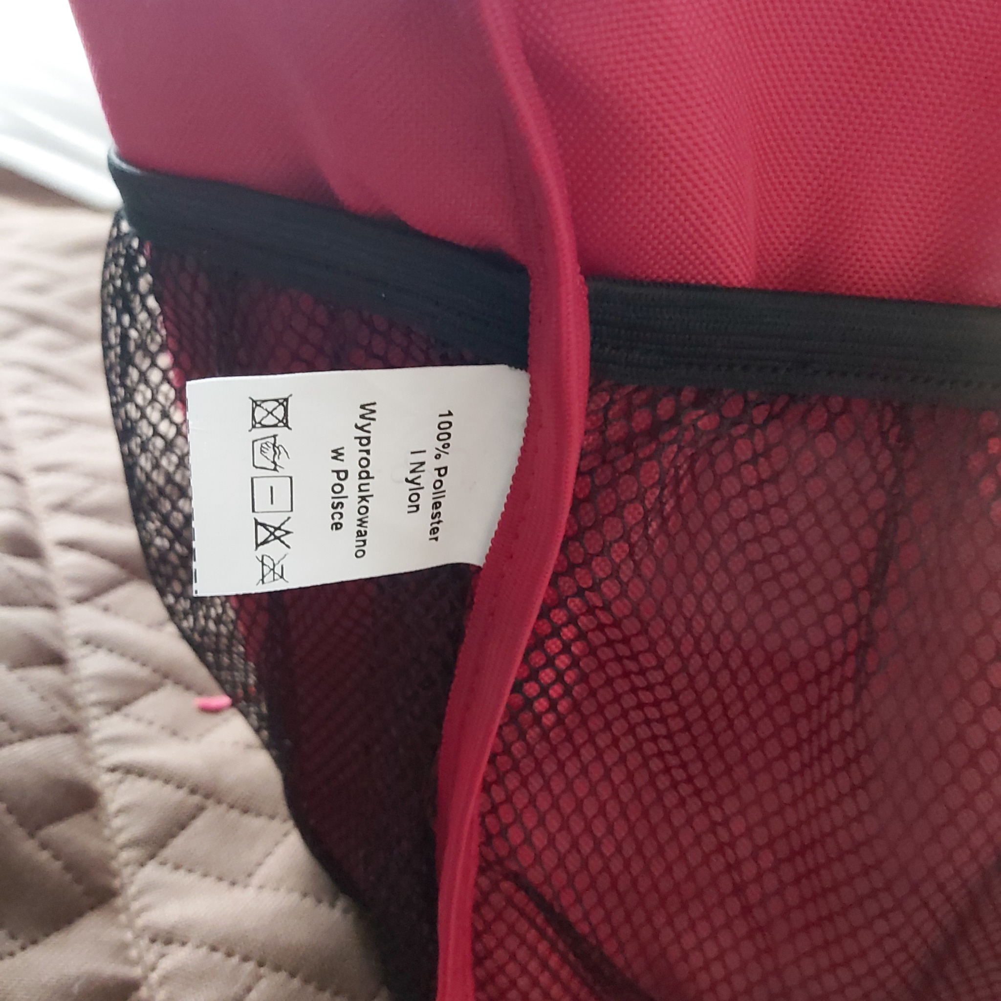 Czerwony plecak z siatkową kieszenią boczną i wszytą metką z informacją o składzie: 100% poliester i nylon, wyprodukowano w Polsce, oraz symbolami prania.