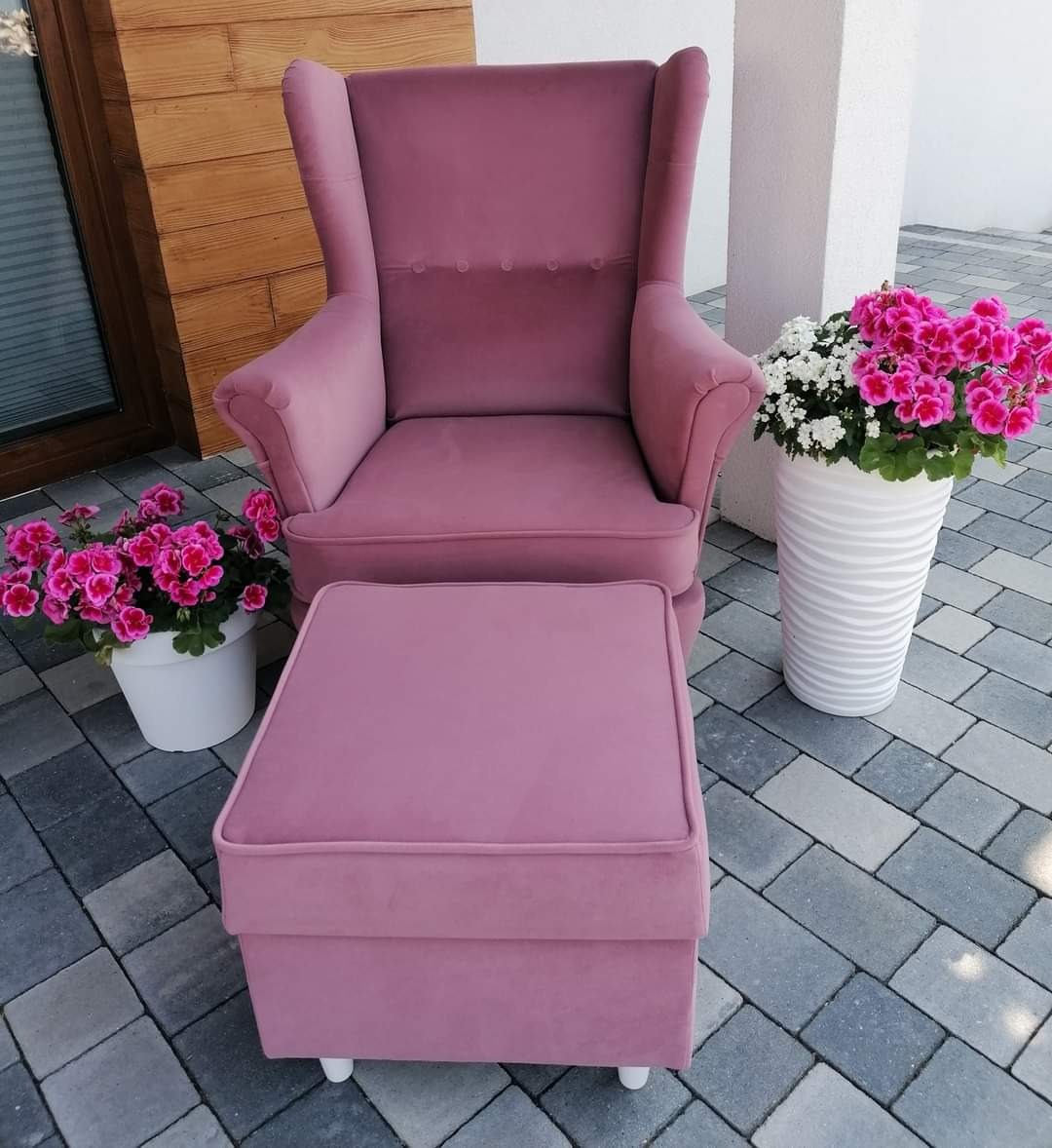 Różowy, welurowy fotel typu uszak z podnóżkiem na zewnątrz, obok donic z kwiatami, na tle drewnianej ściany i białego filaru.